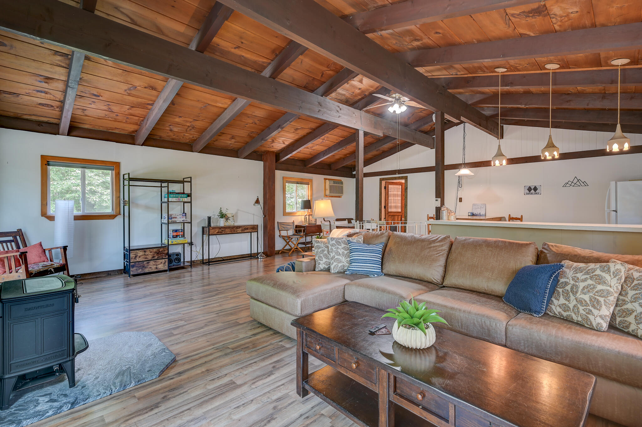 Open living room with vaulted exposed-beam ceiling, sectional sofa, wood stove, and kitchen counter.