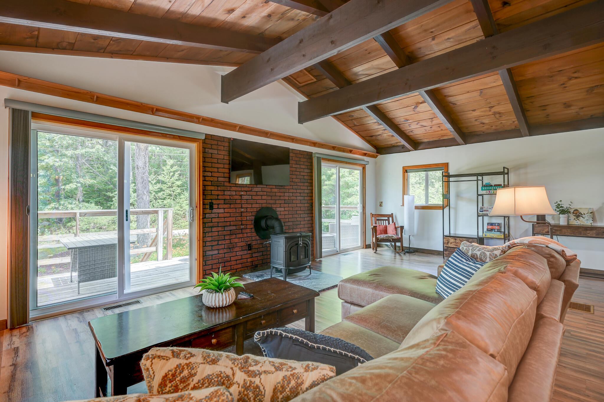 Living room with a vaulted timber ceiling, brick stove wall, and sliding doors to a wooded deck.