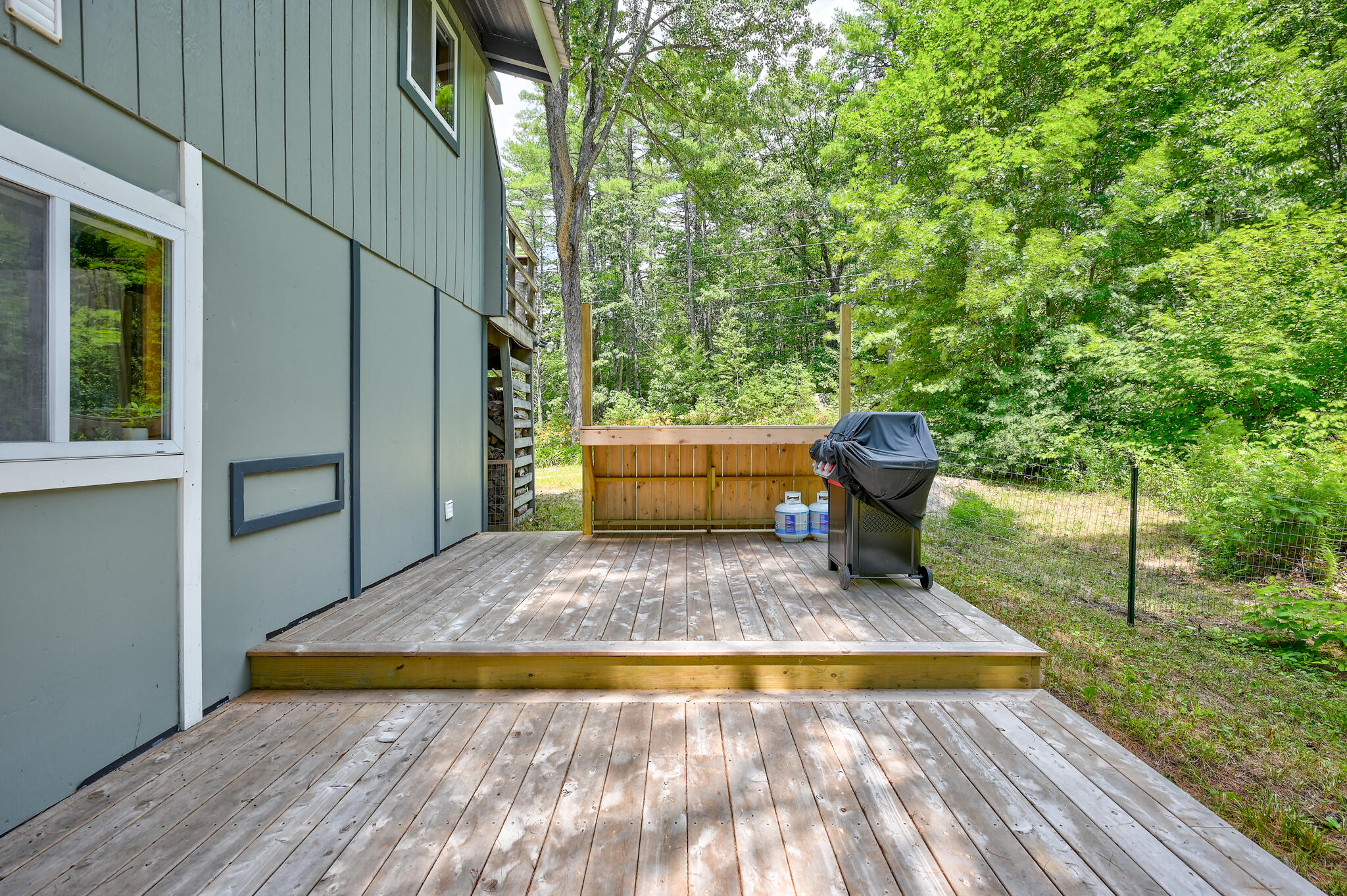 Raised wooden deck beside a gray-sided home, with a covered grill and wooded yard.