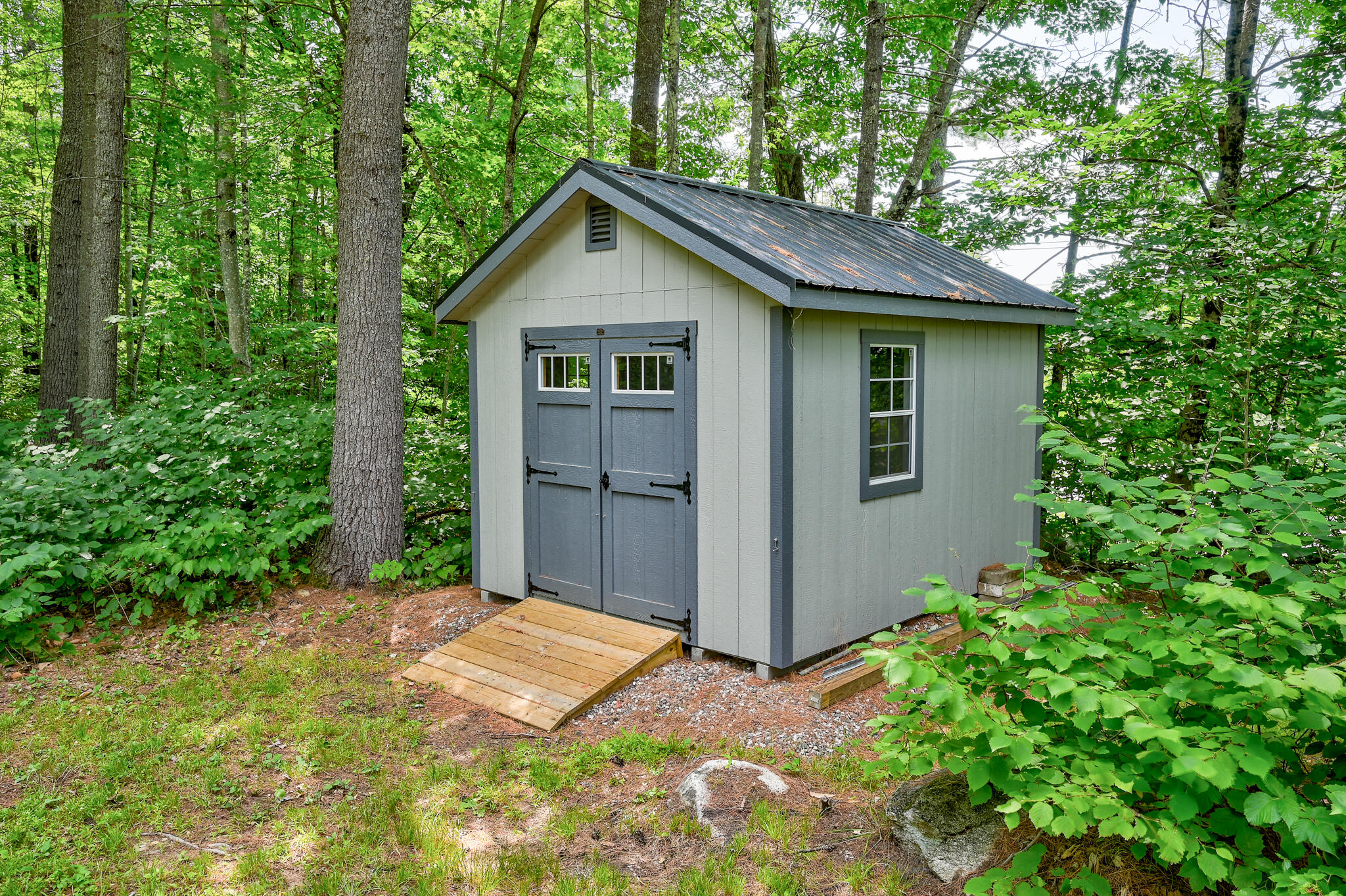 Gray garden shed with double doors and a wooden ramp, surrounded by leafy woods.