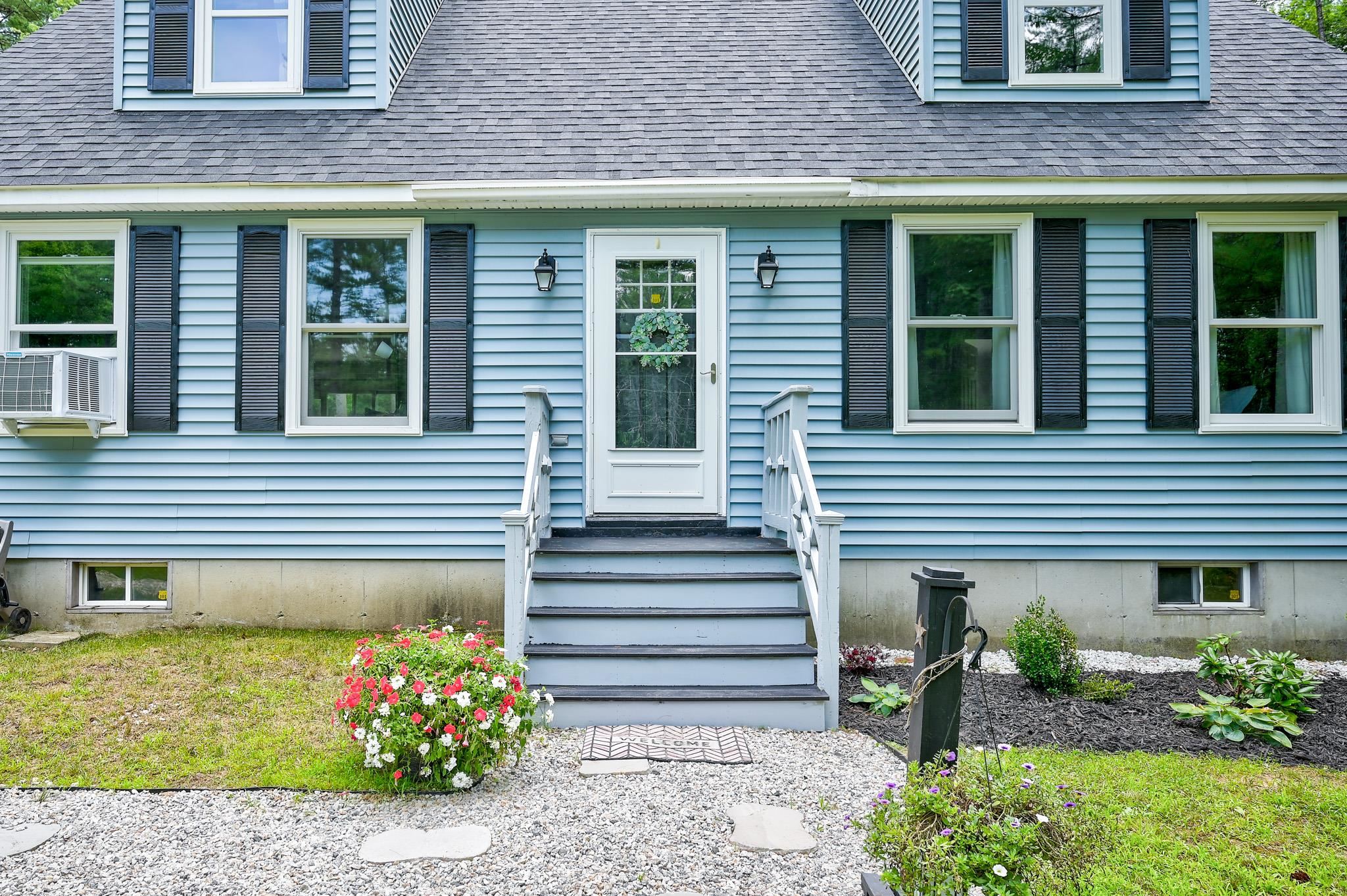 Light blue house entrance with white steps, dark shutters, and flowers beside a gravel path.