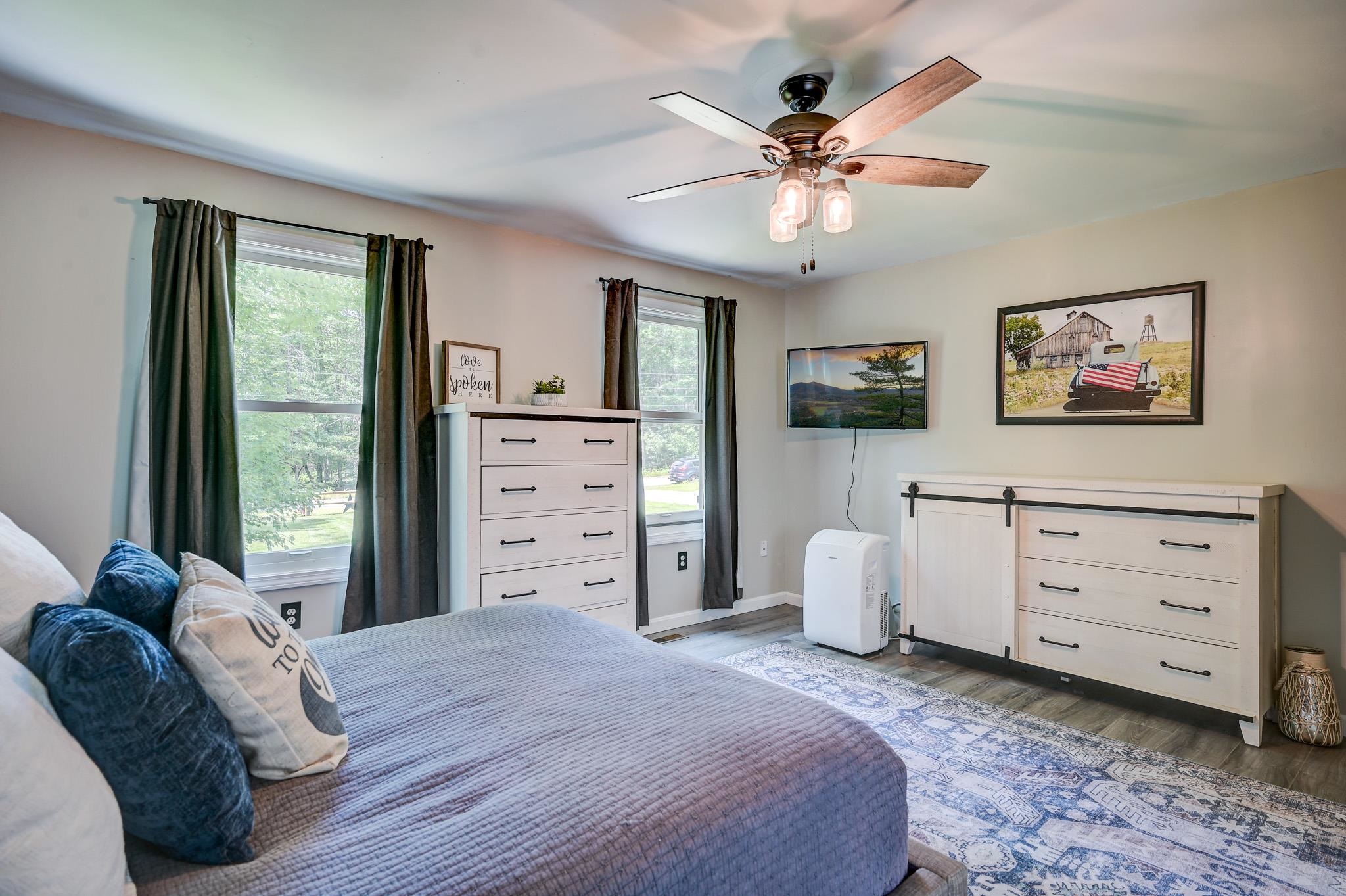 Bedroom with blue bedding, white dressers, two curtained windows, and a ceiling fan.