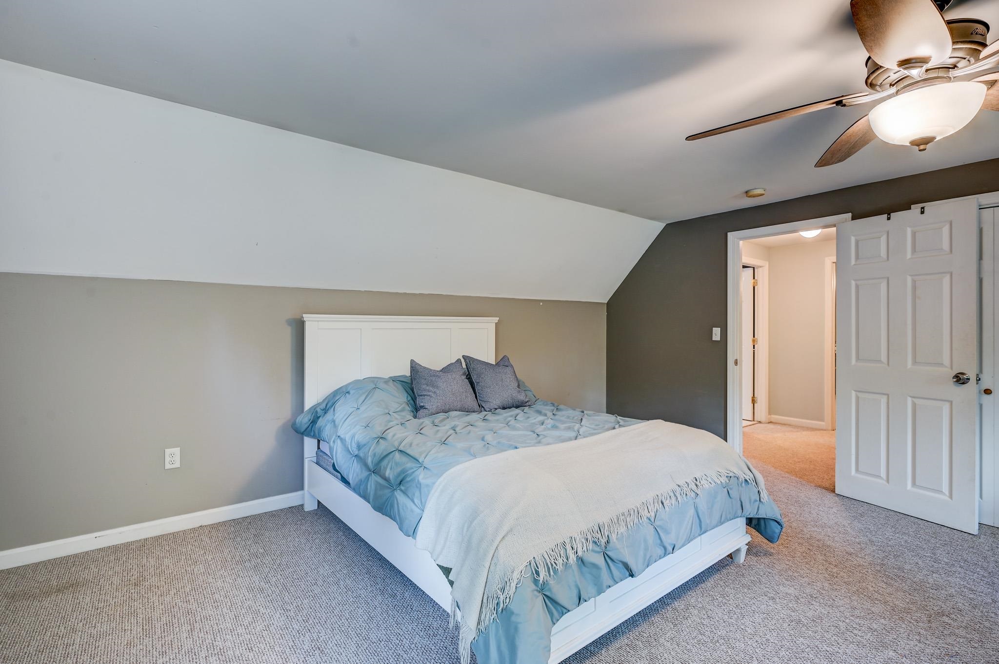 Staged bedroom with a white bed, blue bedding, sloped ceiling, and ceiling fan.
