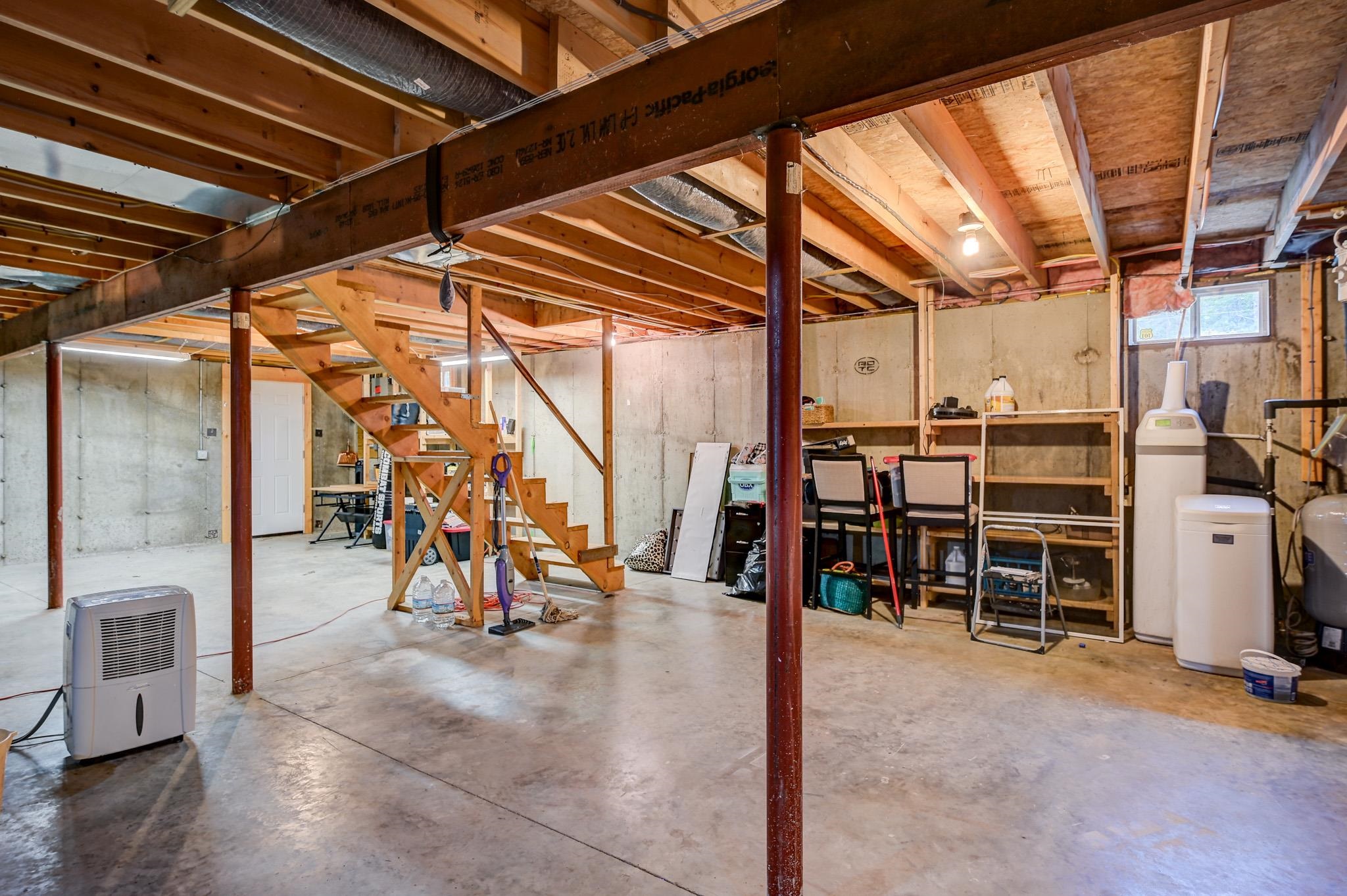 Unfinished basement with exposed ceiling joists, concrete floor, wooden staircase, and utility equipment.