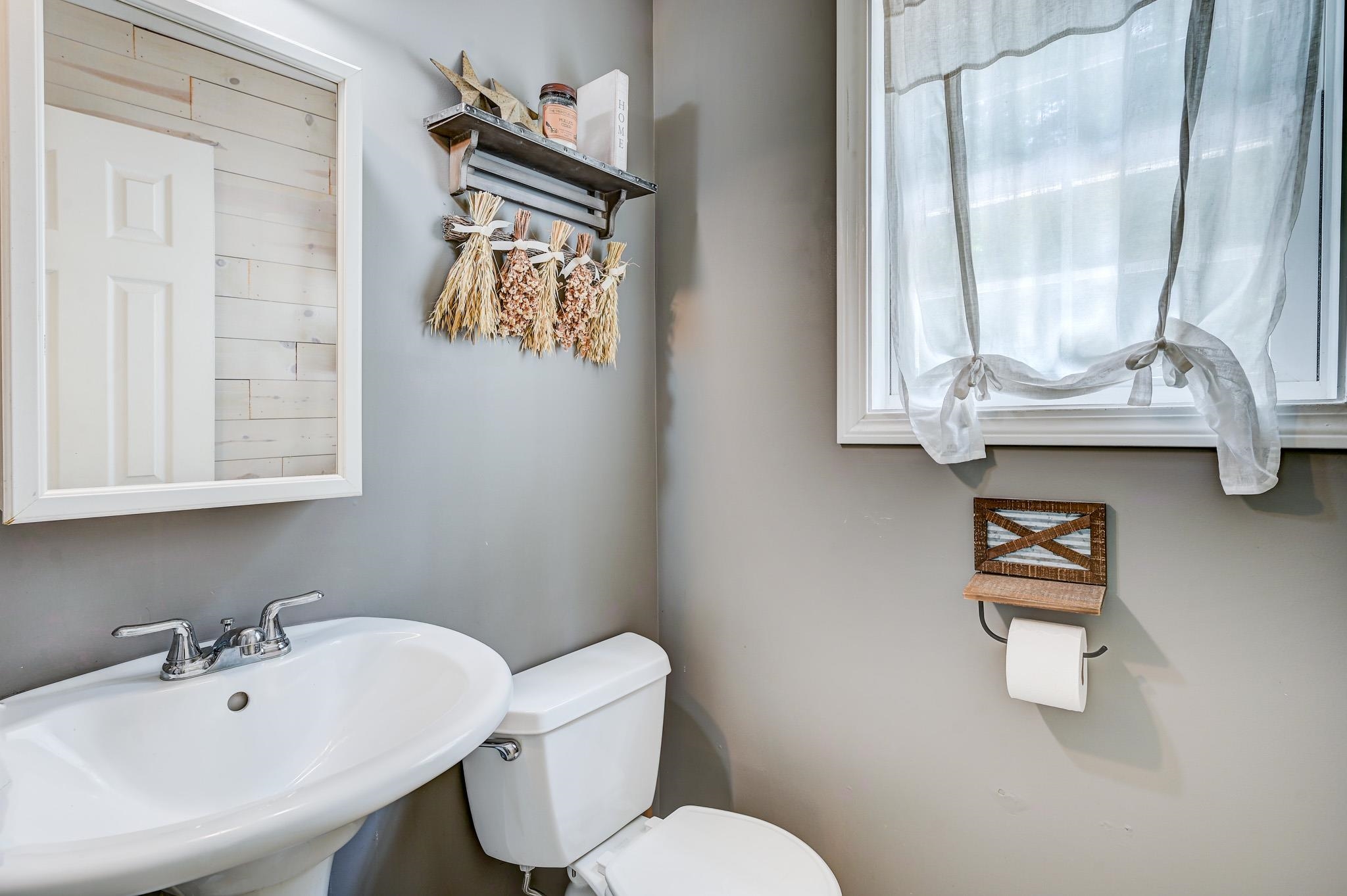 Bathroom with a pedestal sink, toilet, tied-back curtains, and dried flowers on a wall shelf.