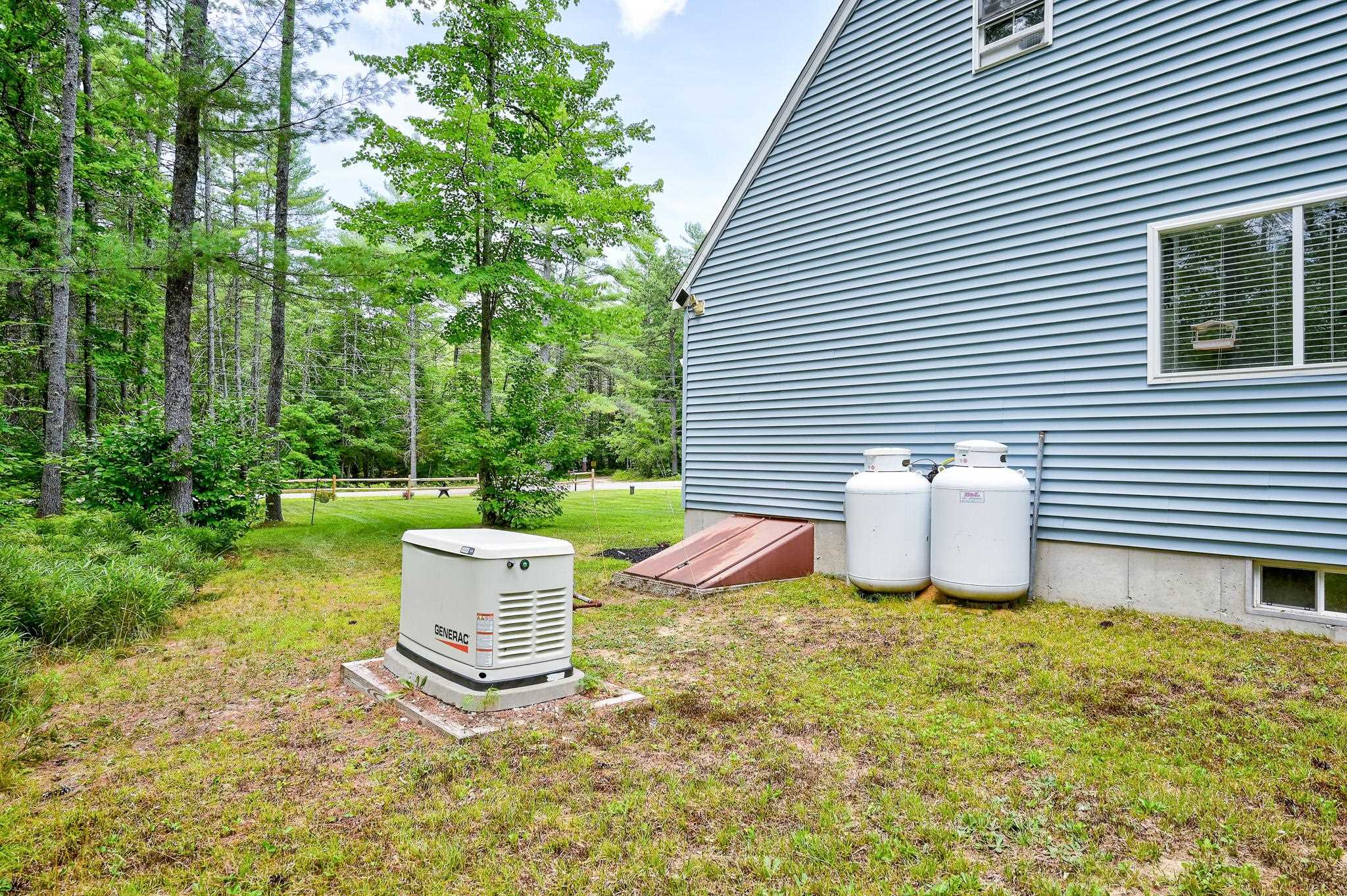 Gray-sided house beside a wooded lawn, with a Generac generator and two propane tanks.