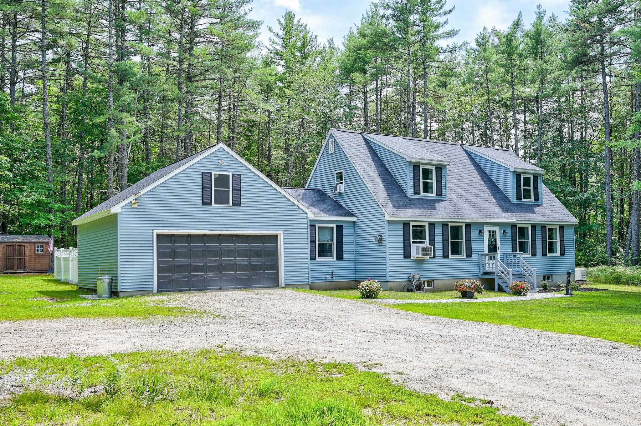 Blue-sided house with a two-car garage and dormer windows, set among trees beside a gravel driveway.