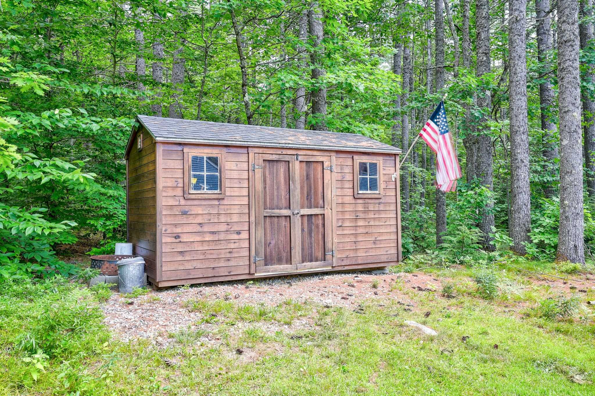 Wooden storage shed with double doors and windows in a wooded clearing, beside an American flag.