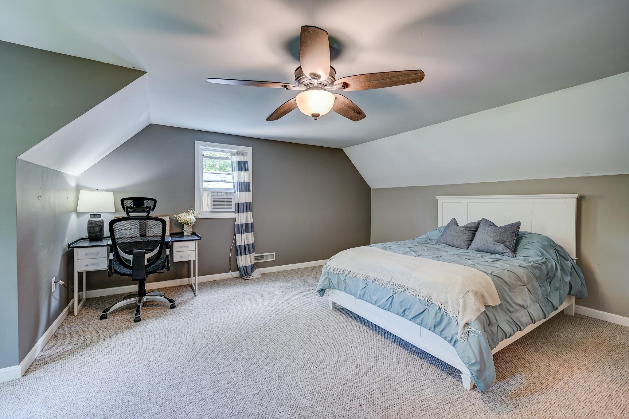 Bedroom with a sloped ceiling, blue-gray bed, and desk beside a curtained window.