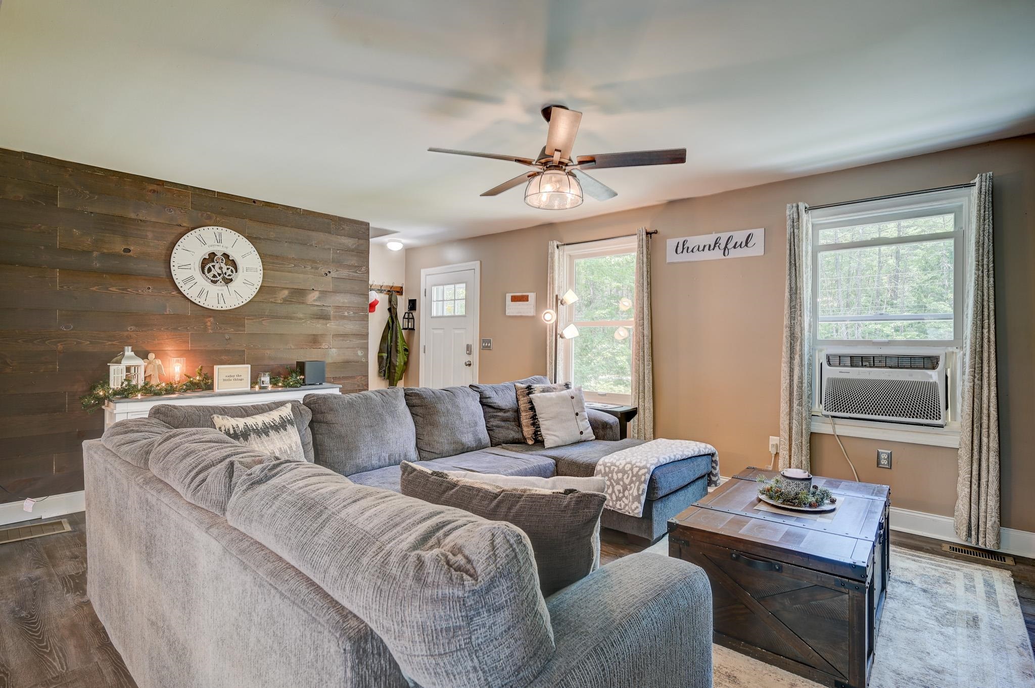Living room with a gray sectional, rustic wood accent wall, large clock, and window air conditioner.