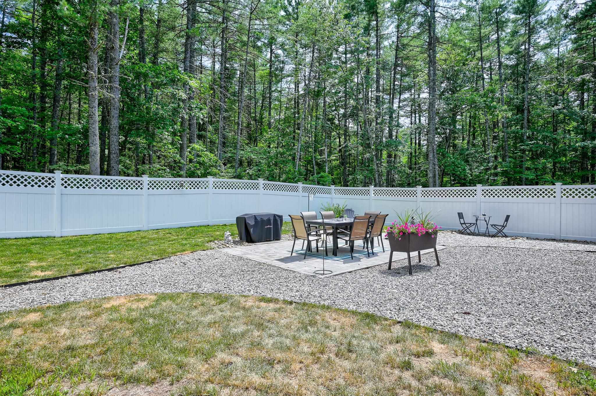 Fenced backyard patio with outdoor dining set, covered grill, and flowers, backed by a wooded area.
