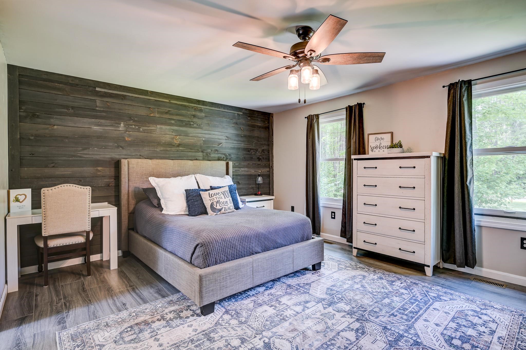 Staged bedroom with a gray bed, dark wood accent wall, white dresser, and blue patterned rug.