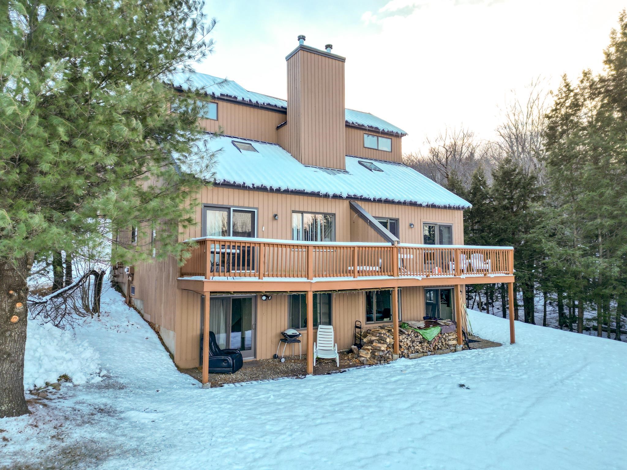 Multi-level tan house with a broad deck, snow-covered roof, and wooded surroundings.