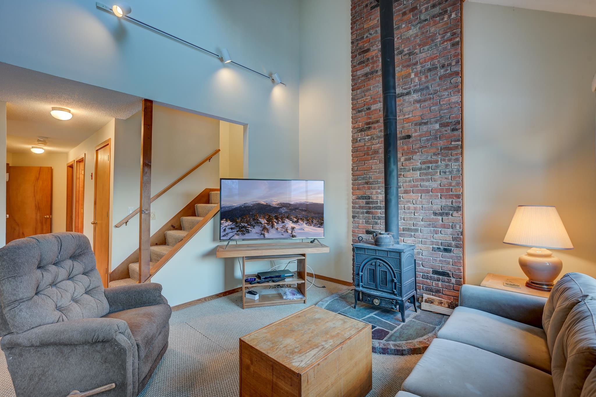 Living room with a brick chimney, wood stove, vaulted ceiling, and staircase.