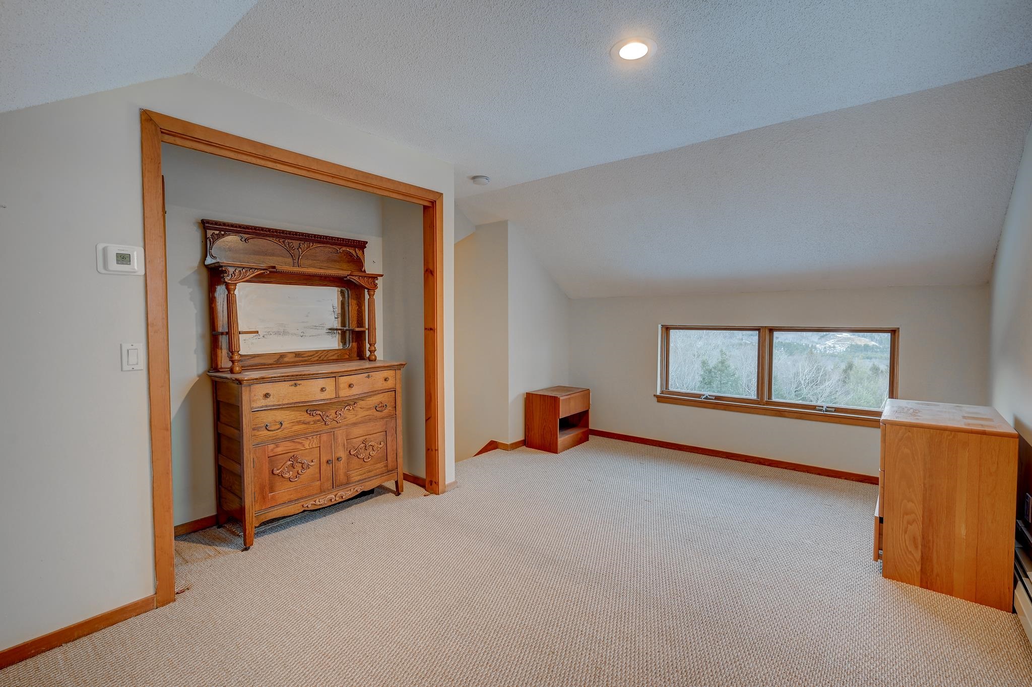 Upstairs bedroom with a sloped ceiling, carved wooden dresser, and windows overlooking trees.