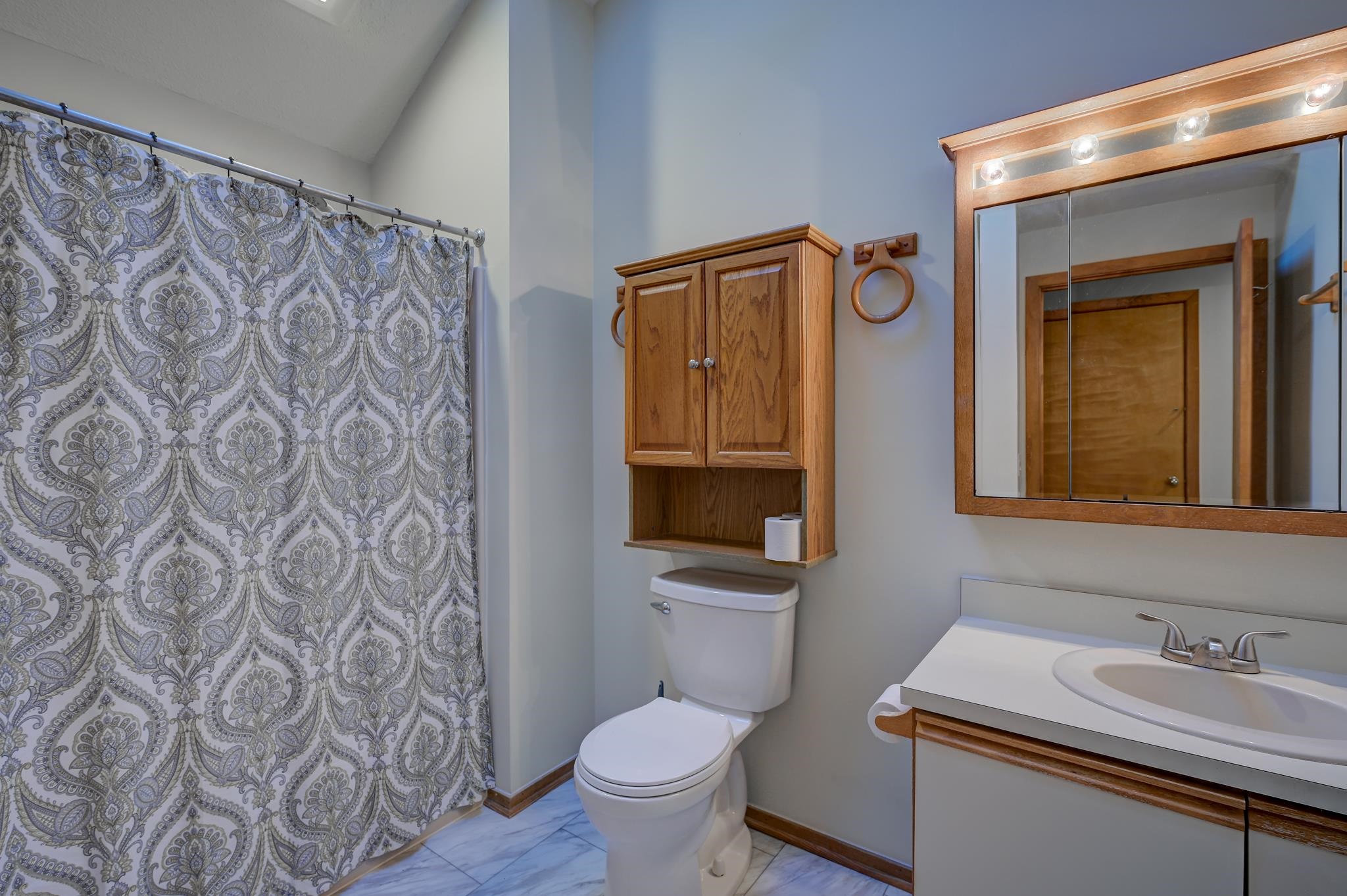Bathroom with patterned shower curtain, wooden storage cabinet, and illuminated mirrored vanity.