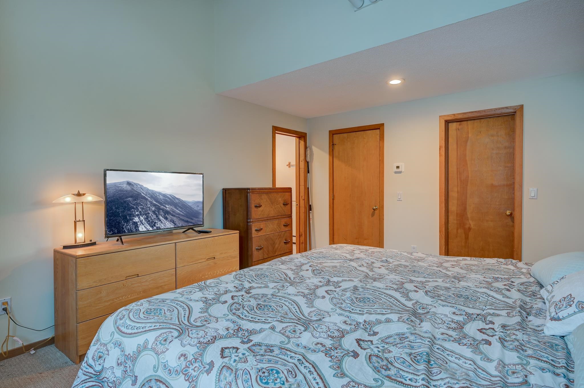 Furnished bedroom with a paisley bedspread, wooden dressers, and a television showing a snowy mountain.