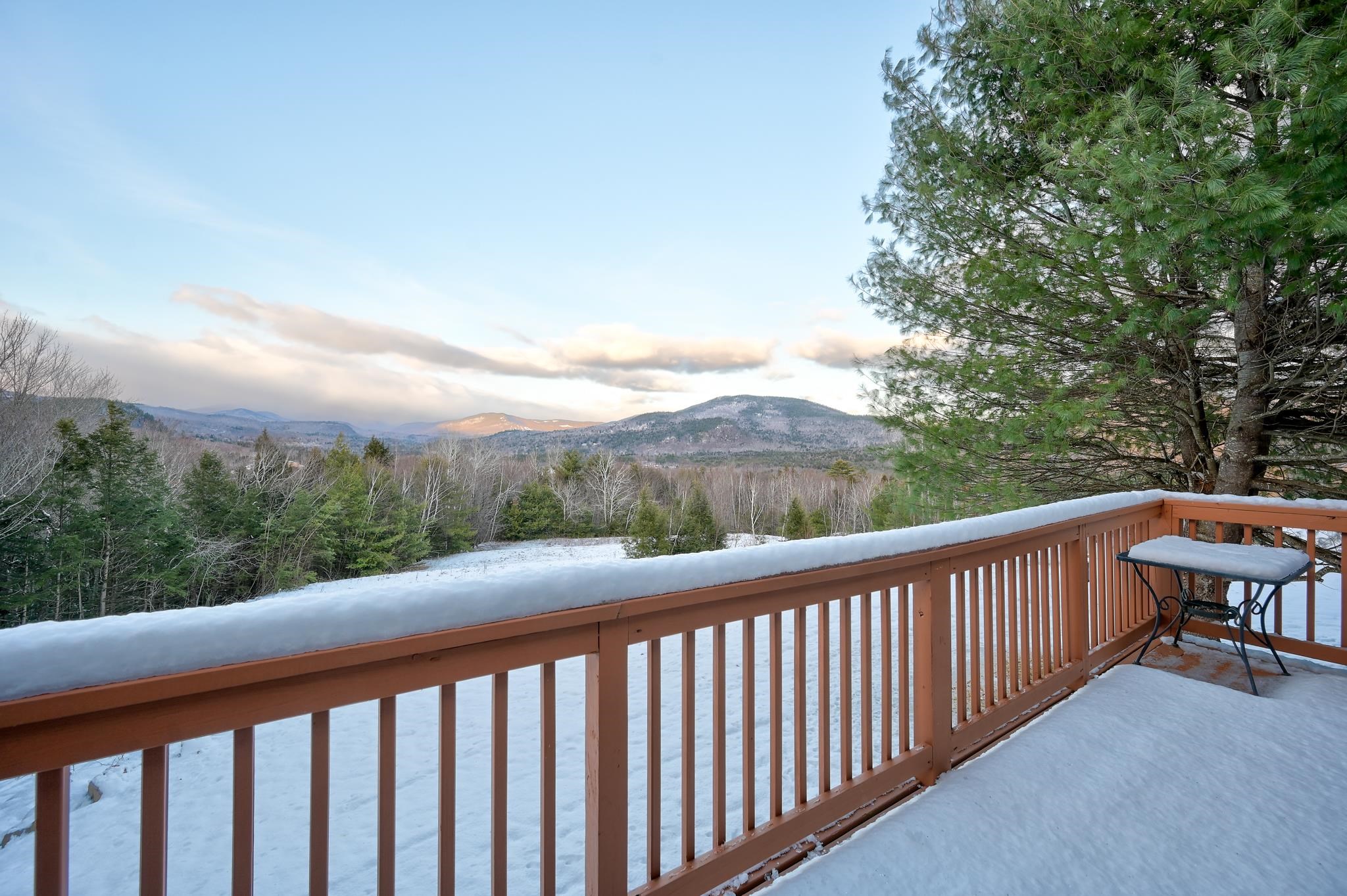 Snow-covered deck overlooking forested mountains, with a pine tree and small table.
