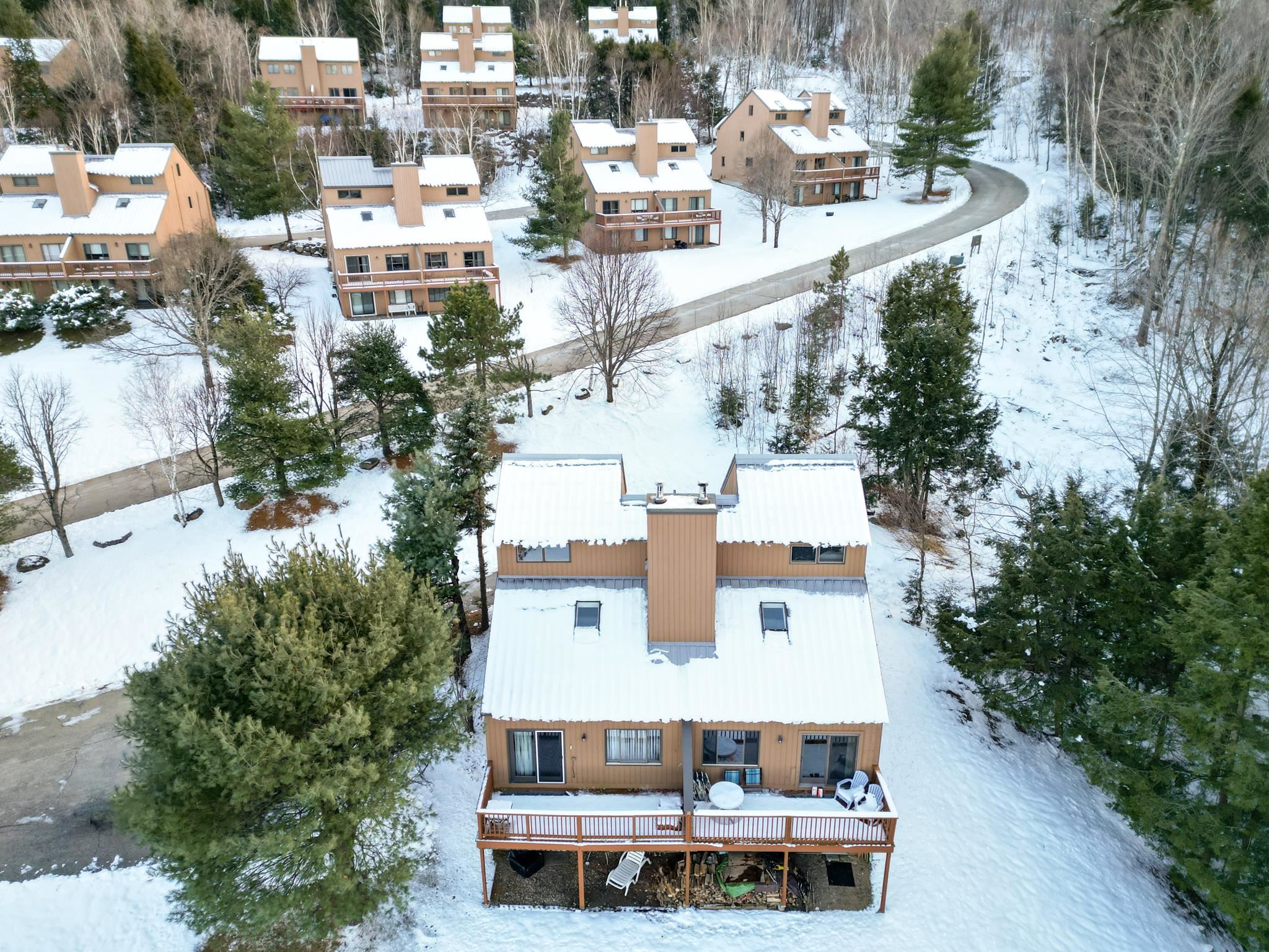 Aerial view of a tan home with a raised deck among snow-covered trees and neighboring homes.