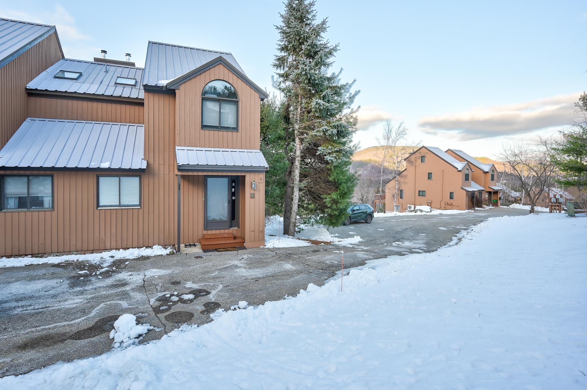 Brown multi-level home with a metal roof beside a snowy driveway and evergreen tree.