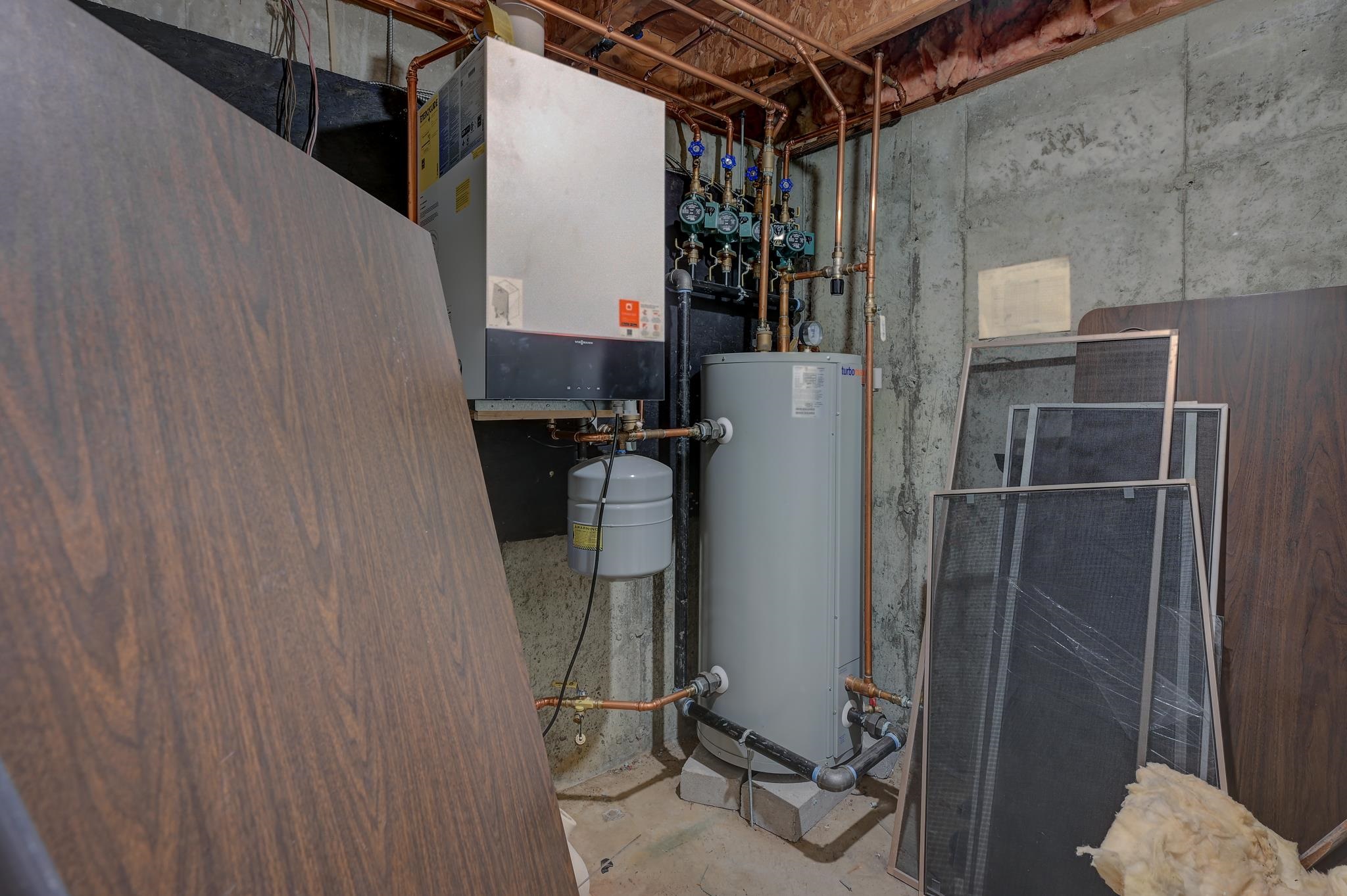 Basement mechanical room with a boiler, hot water tank, exposed copper plumbing, and stored window screens.