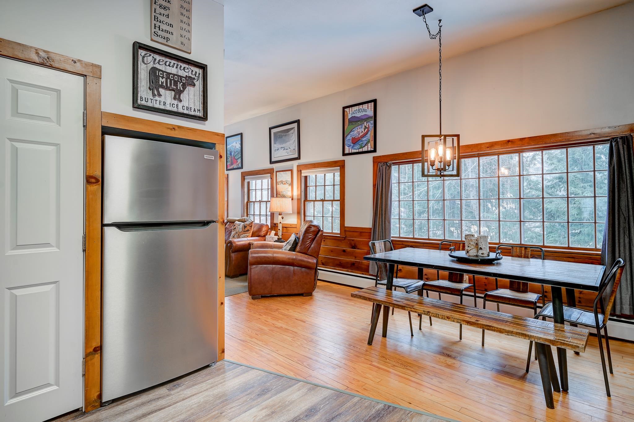 Dining area with a long table beside a large window, wood trim, hardwood floors, and adjoining leather seating.
