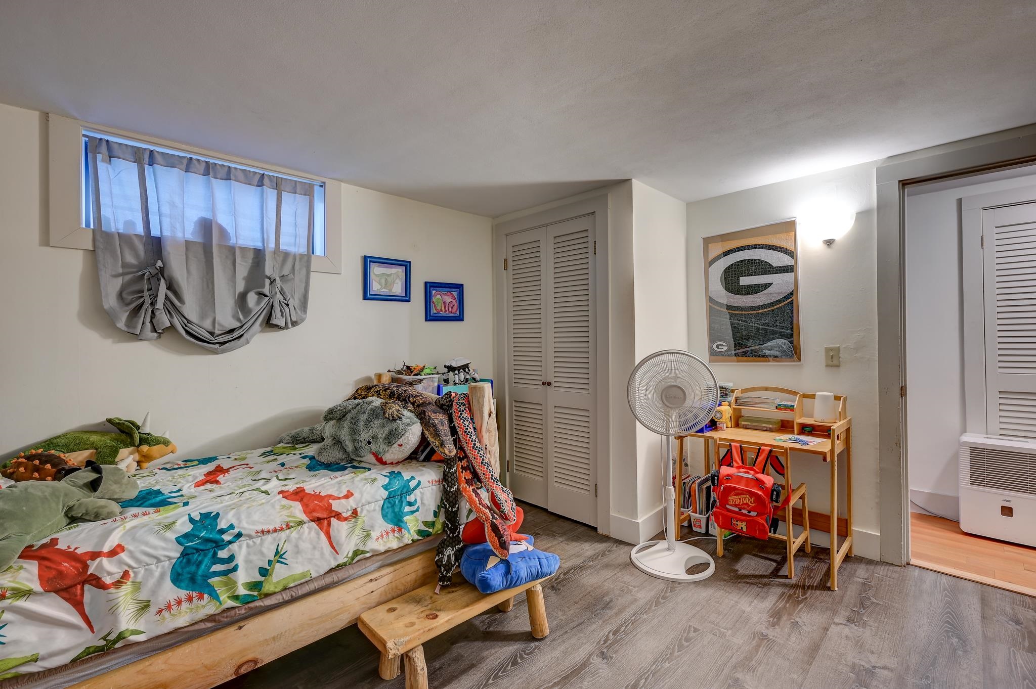 Bedroom with dinosaur-print bedding, plush toys, and a small desk beside the closet.