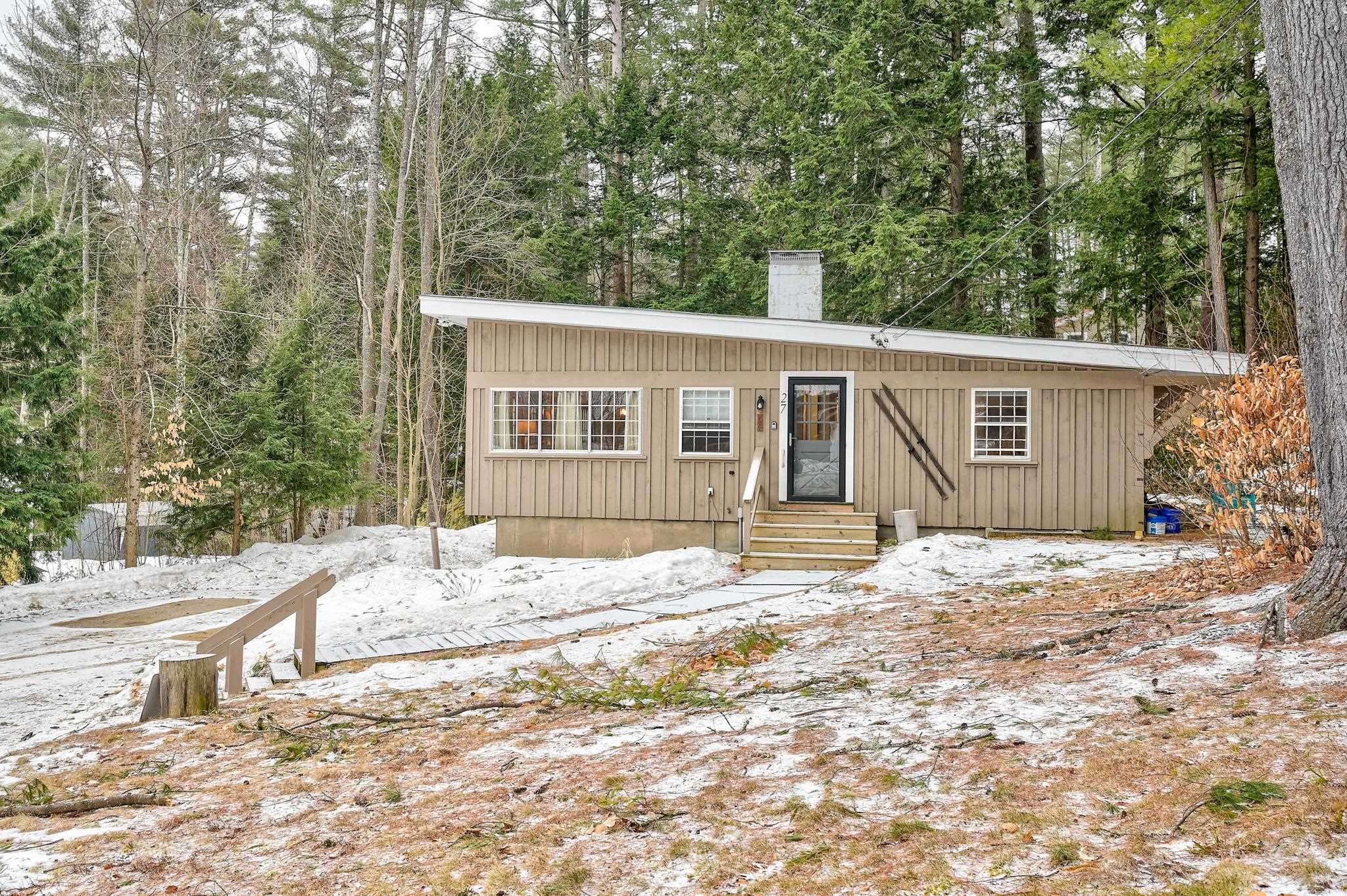 Tan single-story home with a sloping roof and dark entry, surrounded by trees and patchy snow.