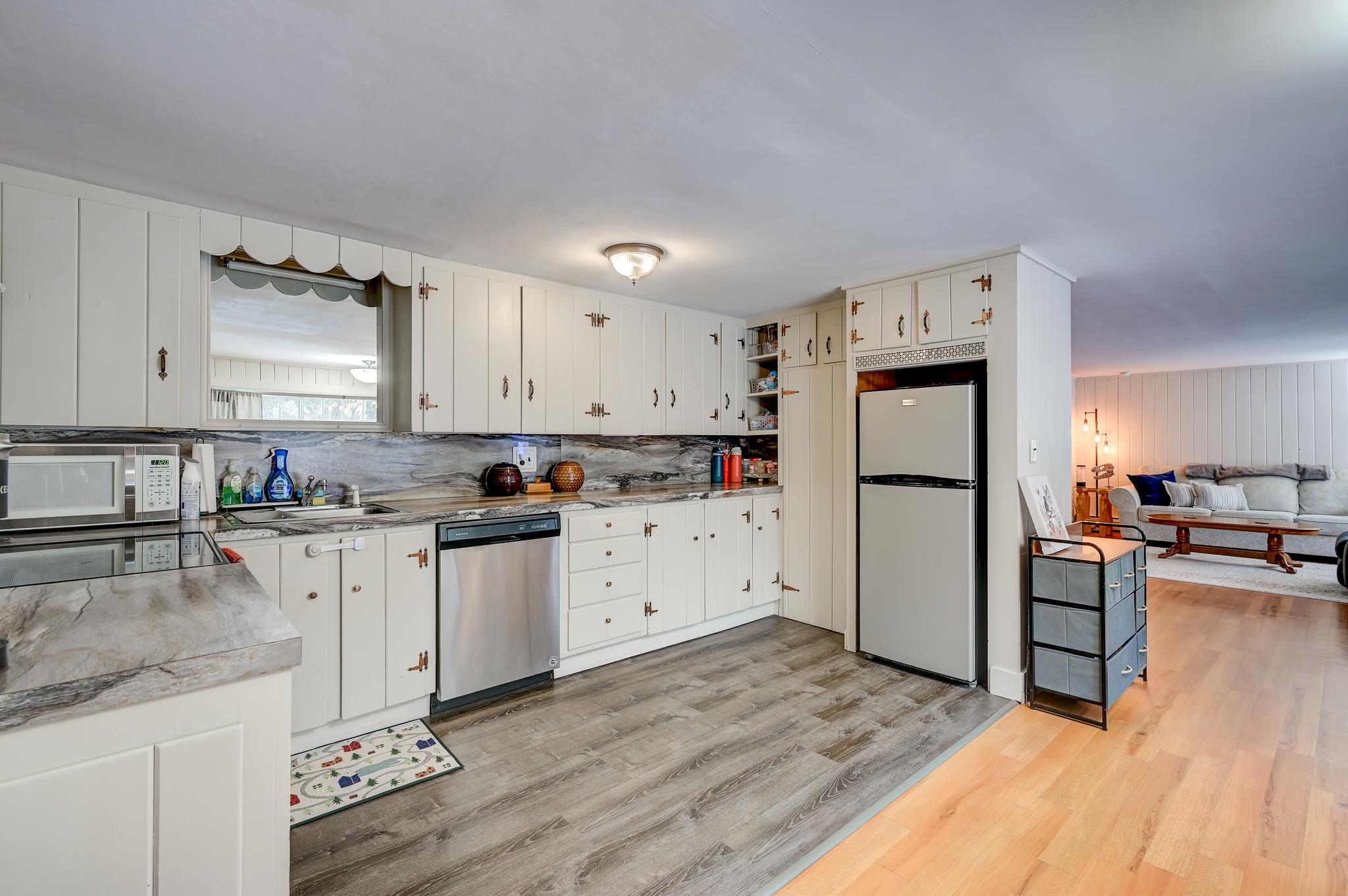 Kitchen with white cabinets and gray stone counters, opening into a living room with a gray sofa.