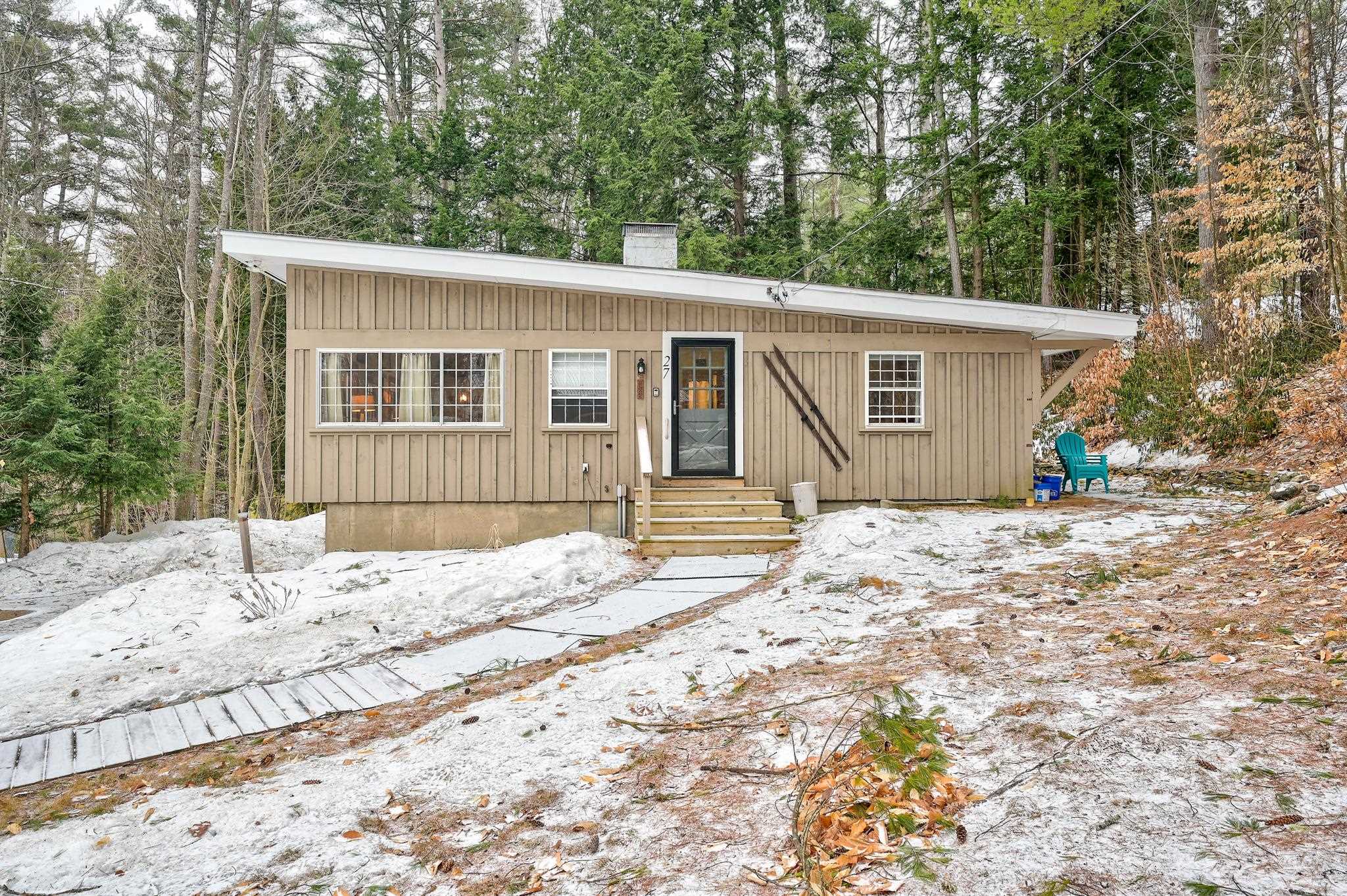 Tan single-story home with a sloping roof and front steps, surrounded by trees and patchy snow.