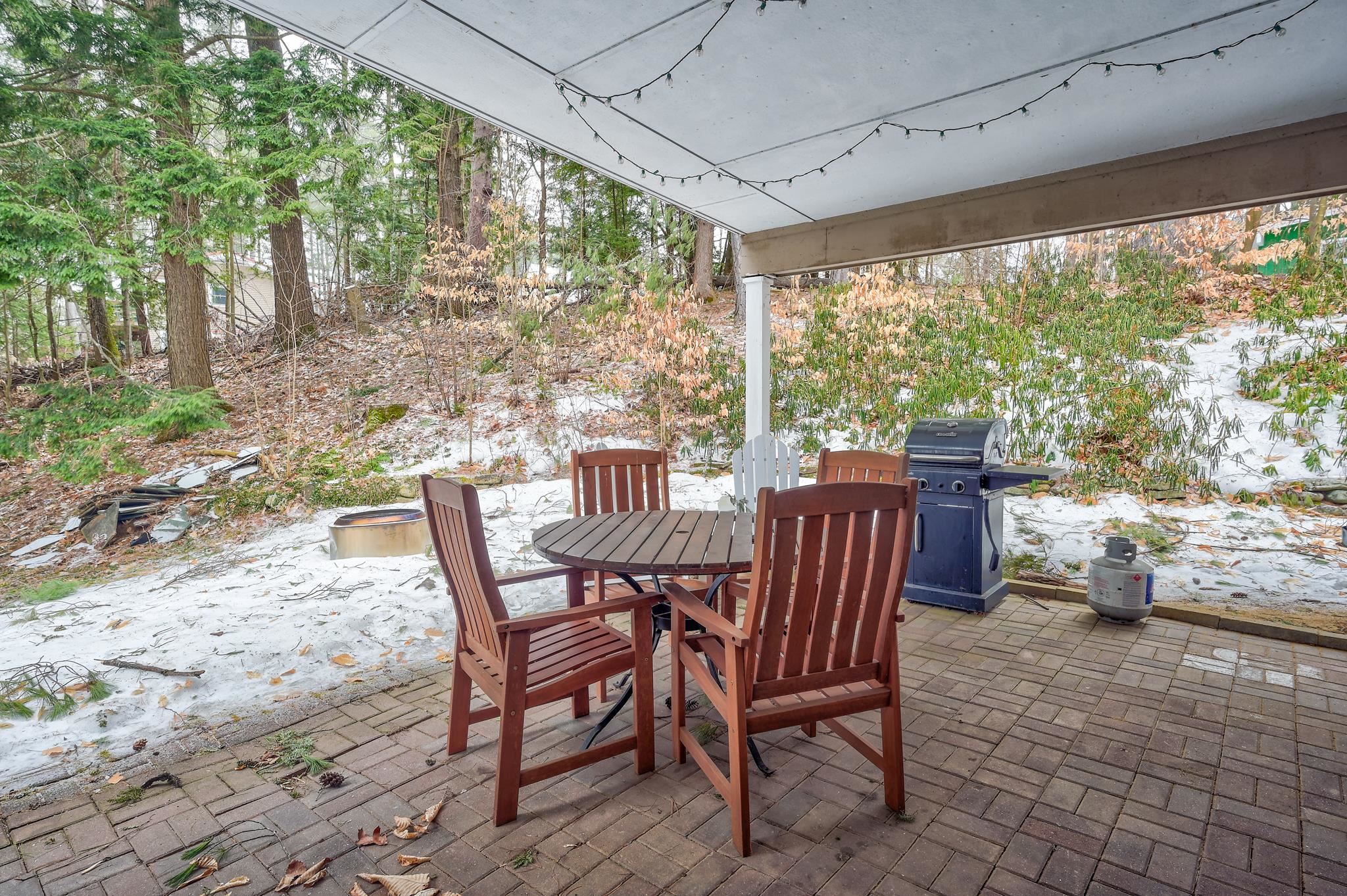 Covered brick patio with wooden dining set and grill beside a lightly snow-covered wooded yard.