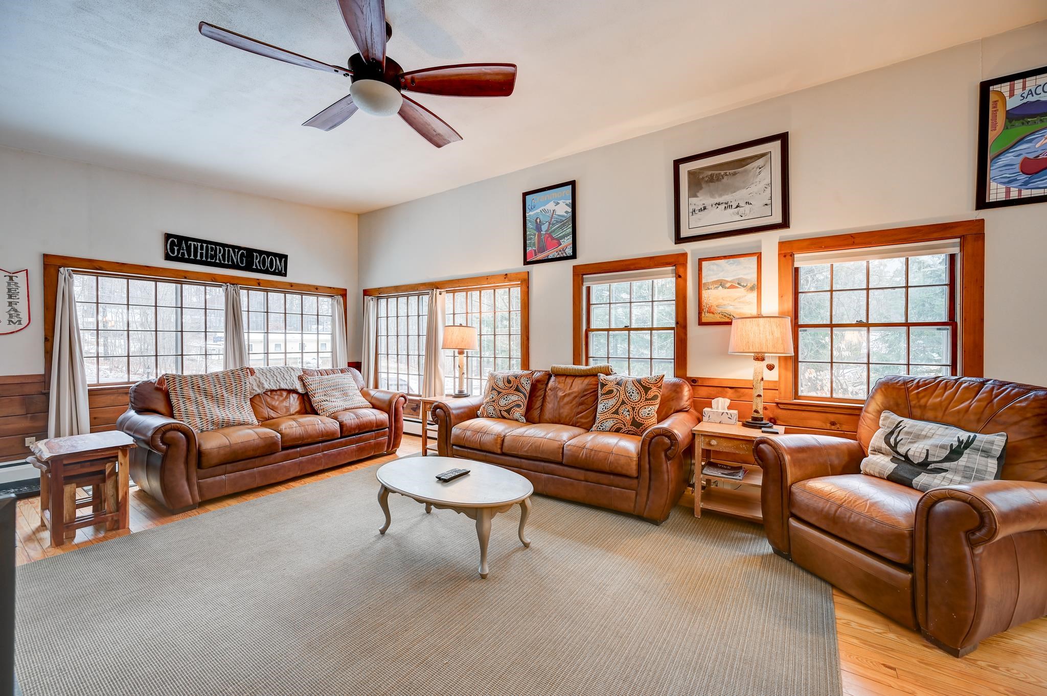 Living room with brown leather seating, wood-trimmed windows, and a sign reading “GATHERING ROOM.”