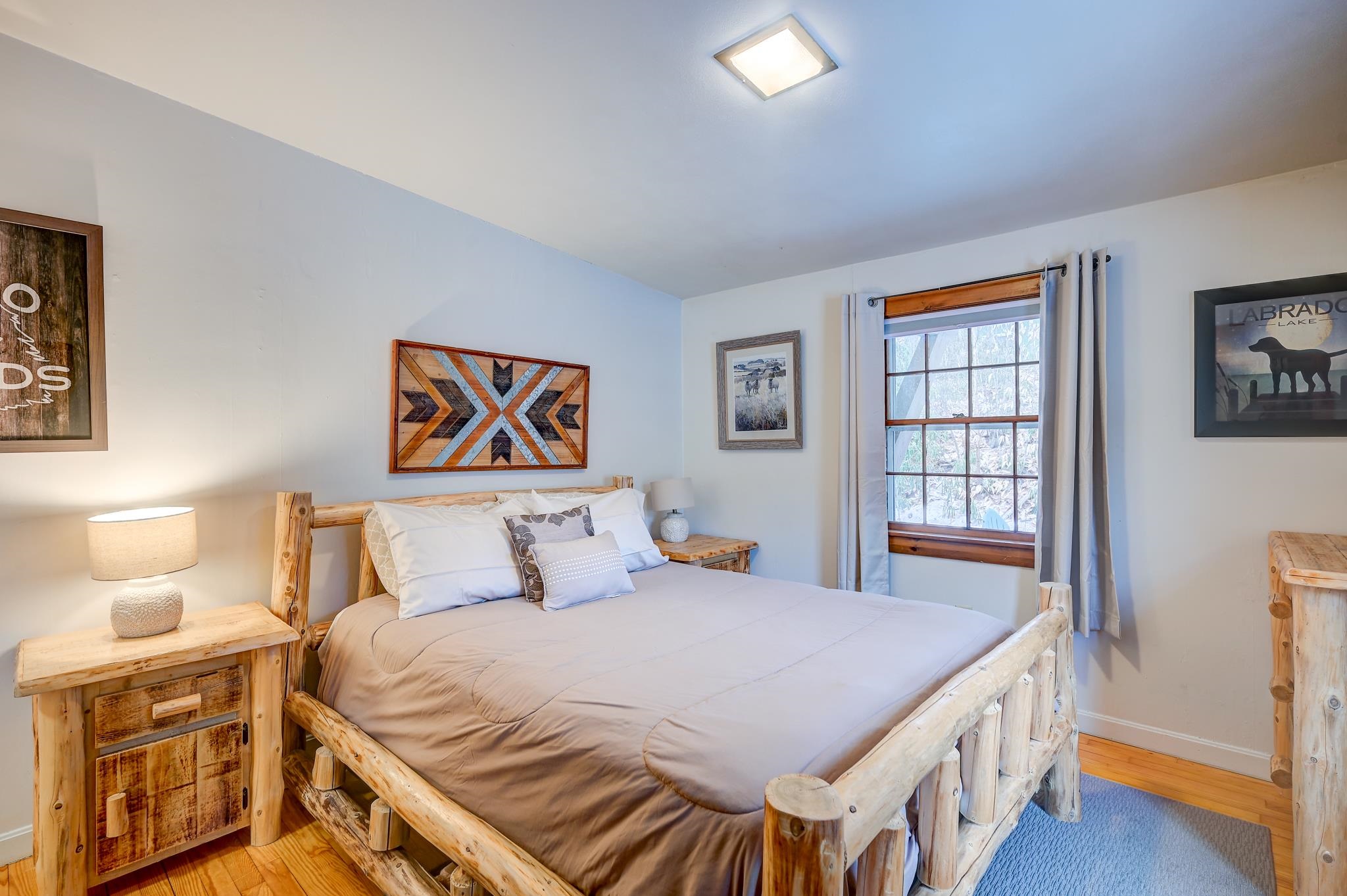 Bedroom with a rustic log bed, neutral bedding, hardwood floors and a curtained window.