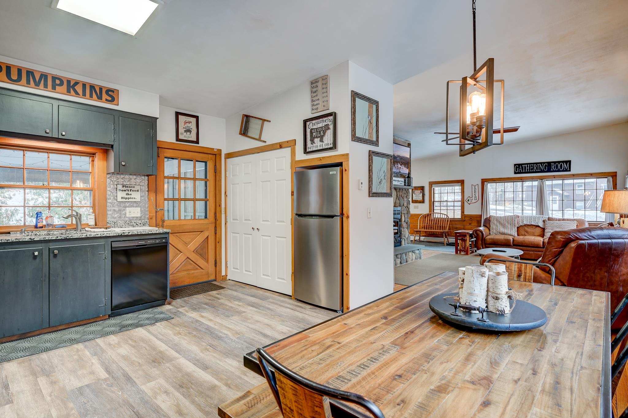 Open kitchen and living area with dark cabinets, a rustic wood dining table, leather sofas, and a stone fireplace.
