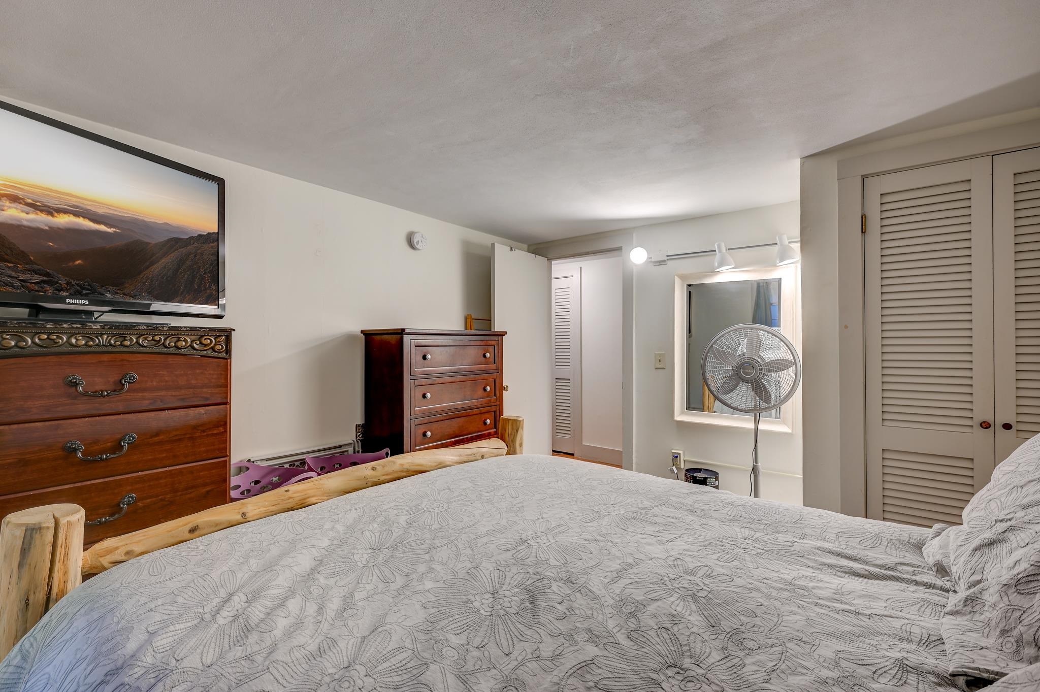 Bedroom with pale floral bedding, rustic wooden dressers, a television, and a standing fan.