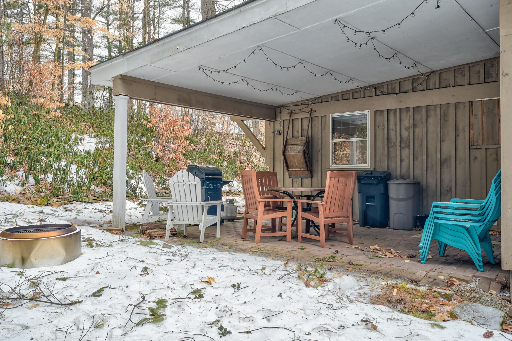 Covered patio with Adirondack chairs, a dining set, grill, and fire pit beside snow-covered woods.
