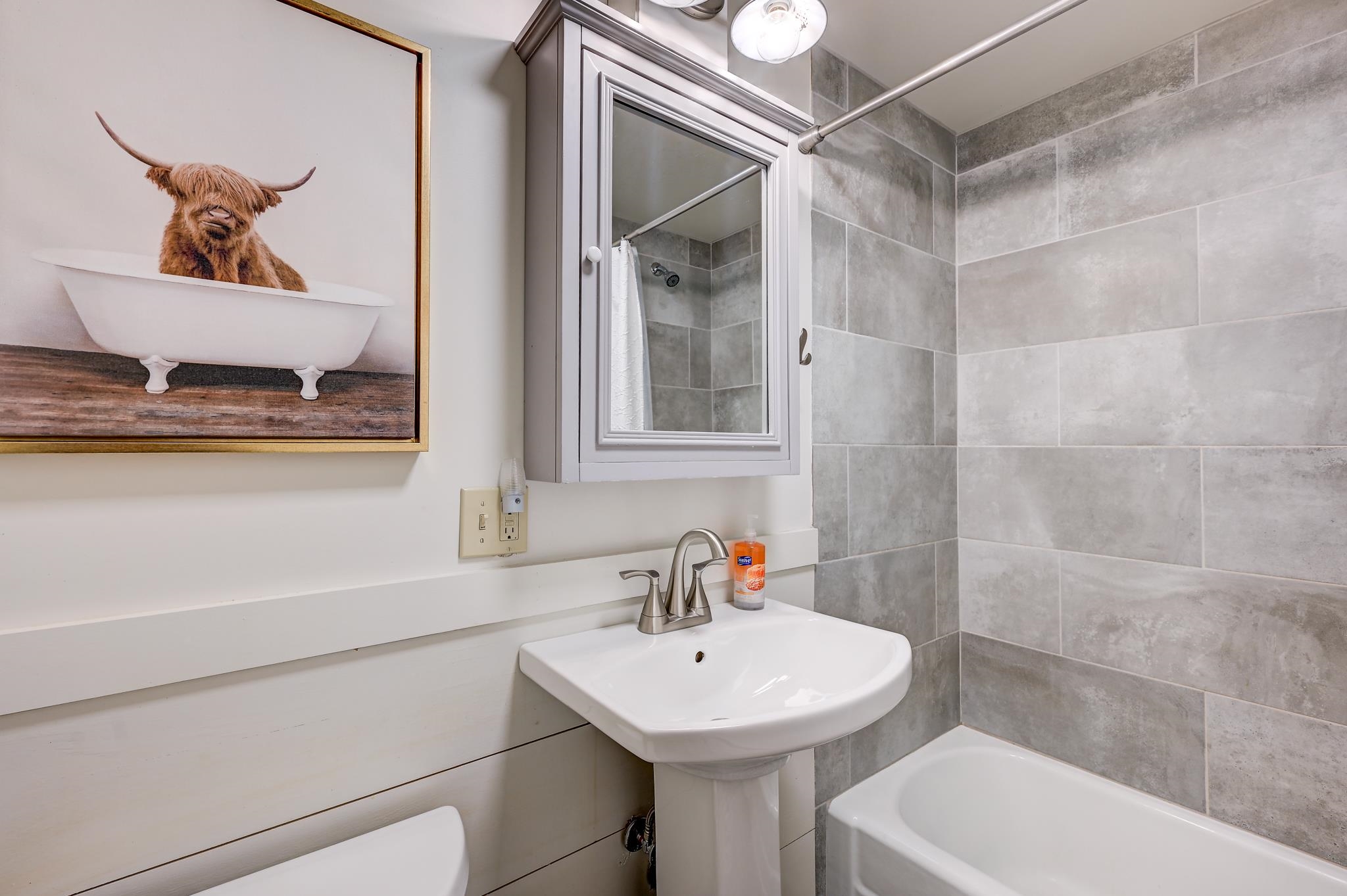 Bathroom with gray tile, a pedestal sink, bathtub, and framed Highland cow artwork.