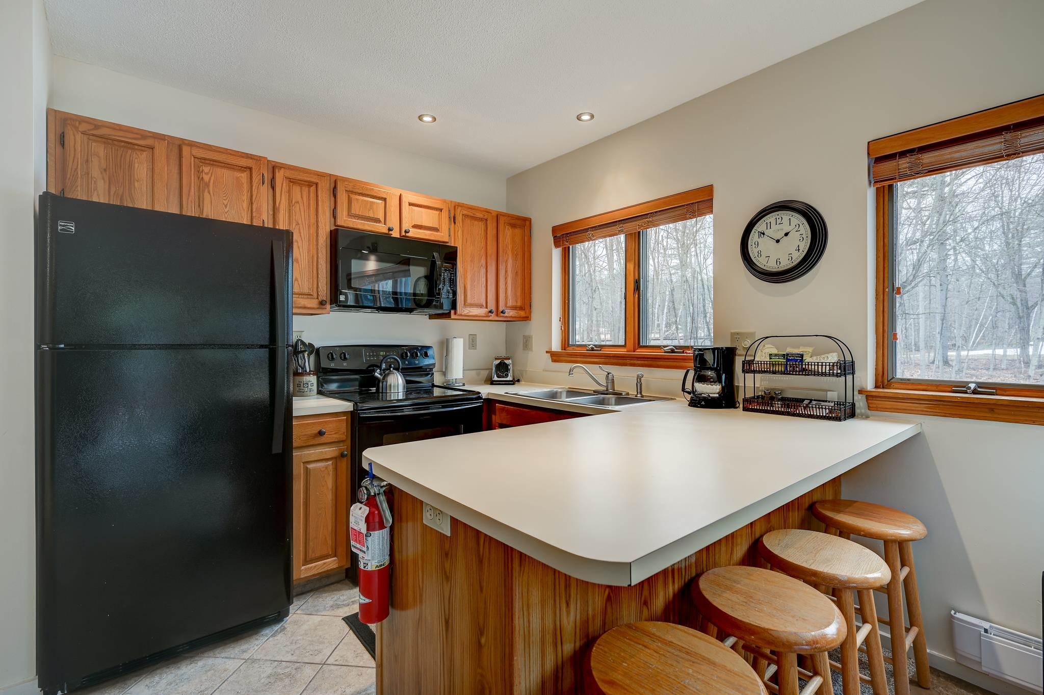 Kitchen with wood cabinets, a white breakfast bar and stools, and windows overlooking snowy woods.