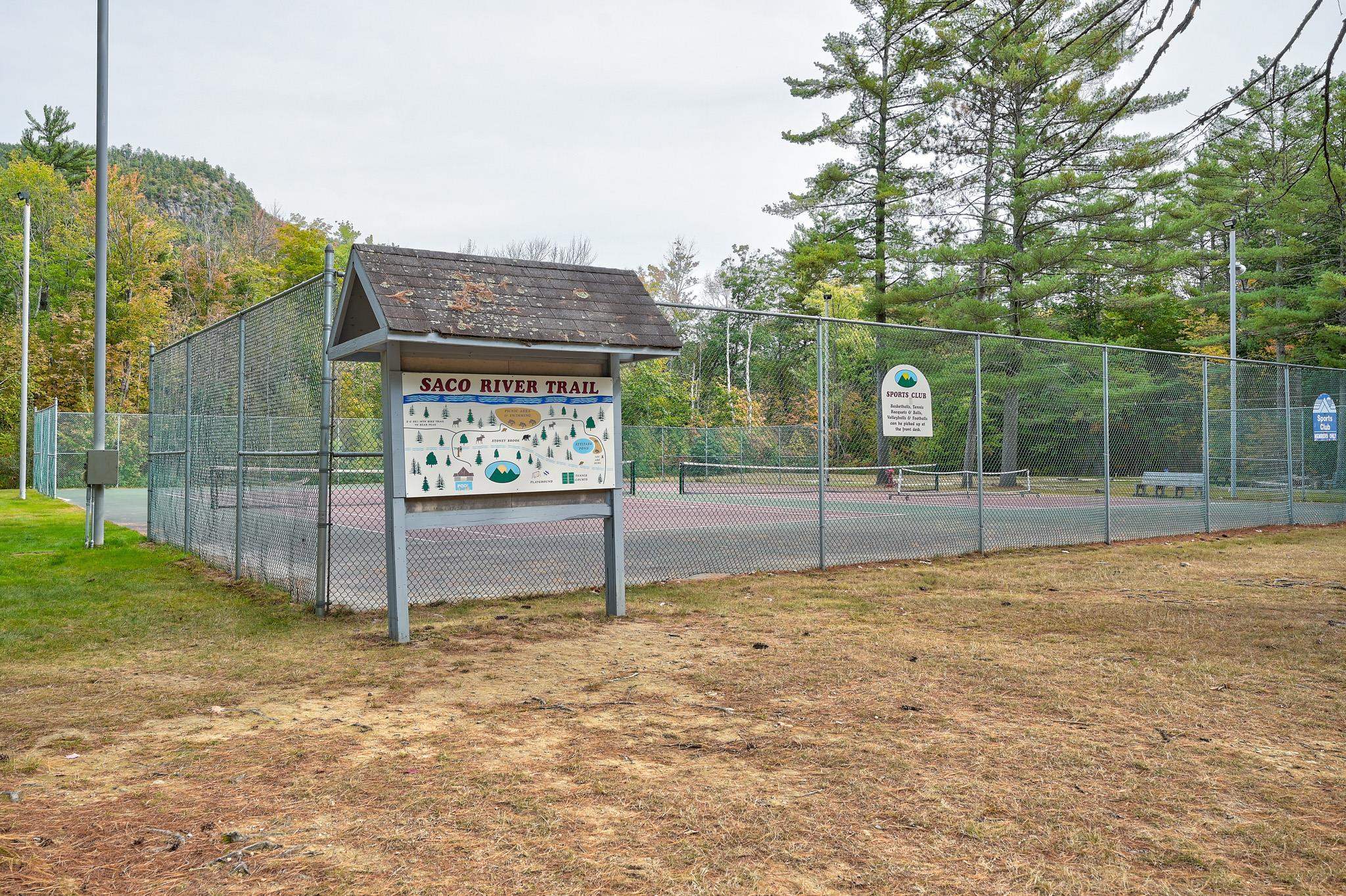 Fenced tennis courts beside a covered Saco River Trail map sign, with a wooded hillside beyond.