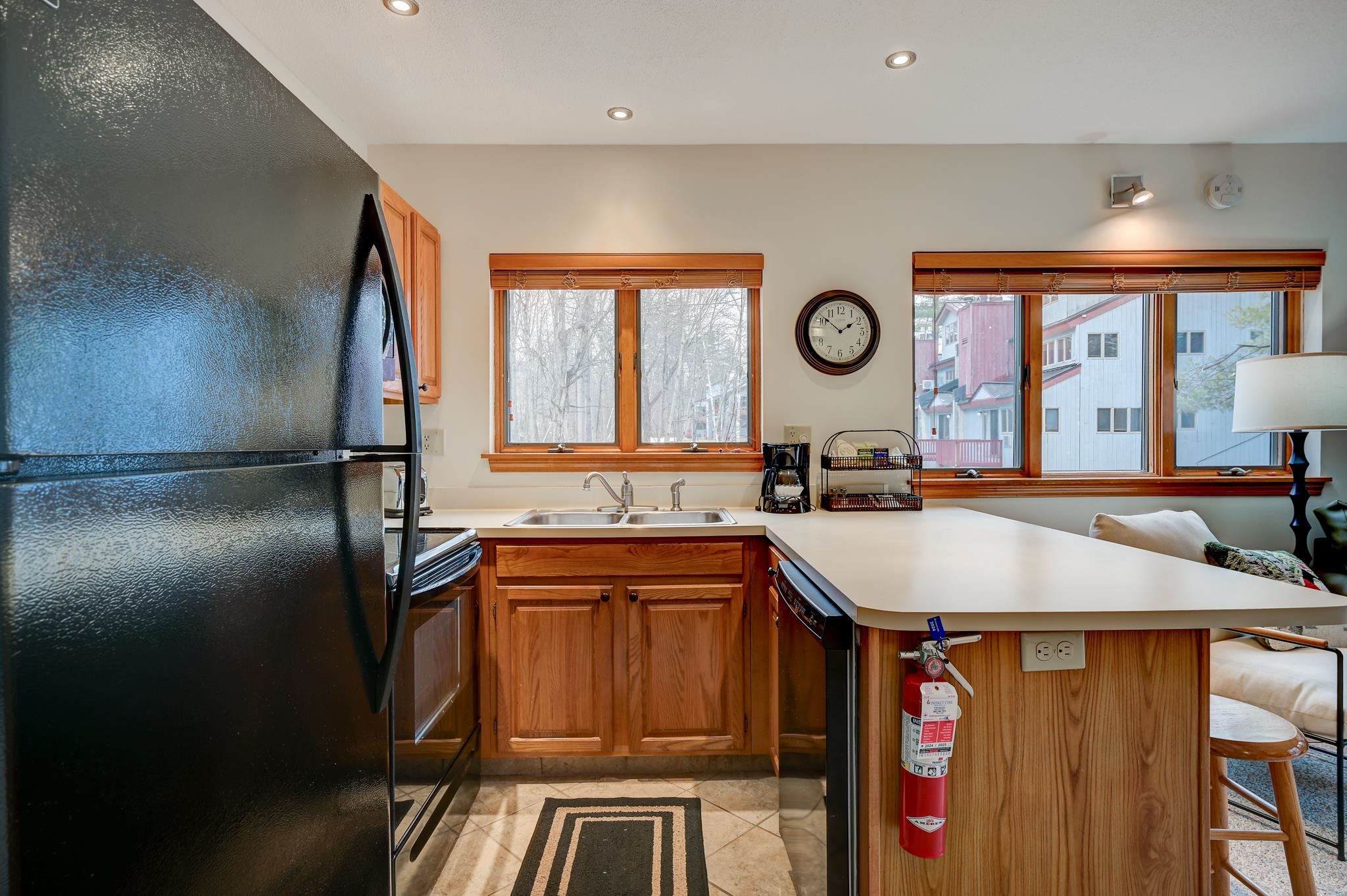 Kitchen with wood cabinets, black refrigerator, double sink, and wide windows overlooking nearby buildings.