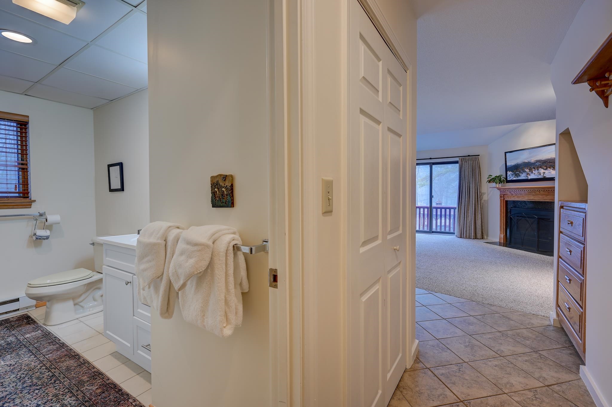 Bathroom with white vanity and towels opens to a living room with a fireplace and balcony doors.