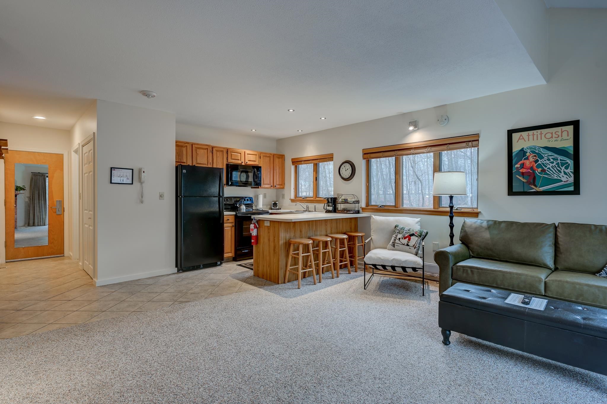 Open-plan condo living room and kitchen with a wood breakfast bar, green sofa, and windows overlooking bare trees.