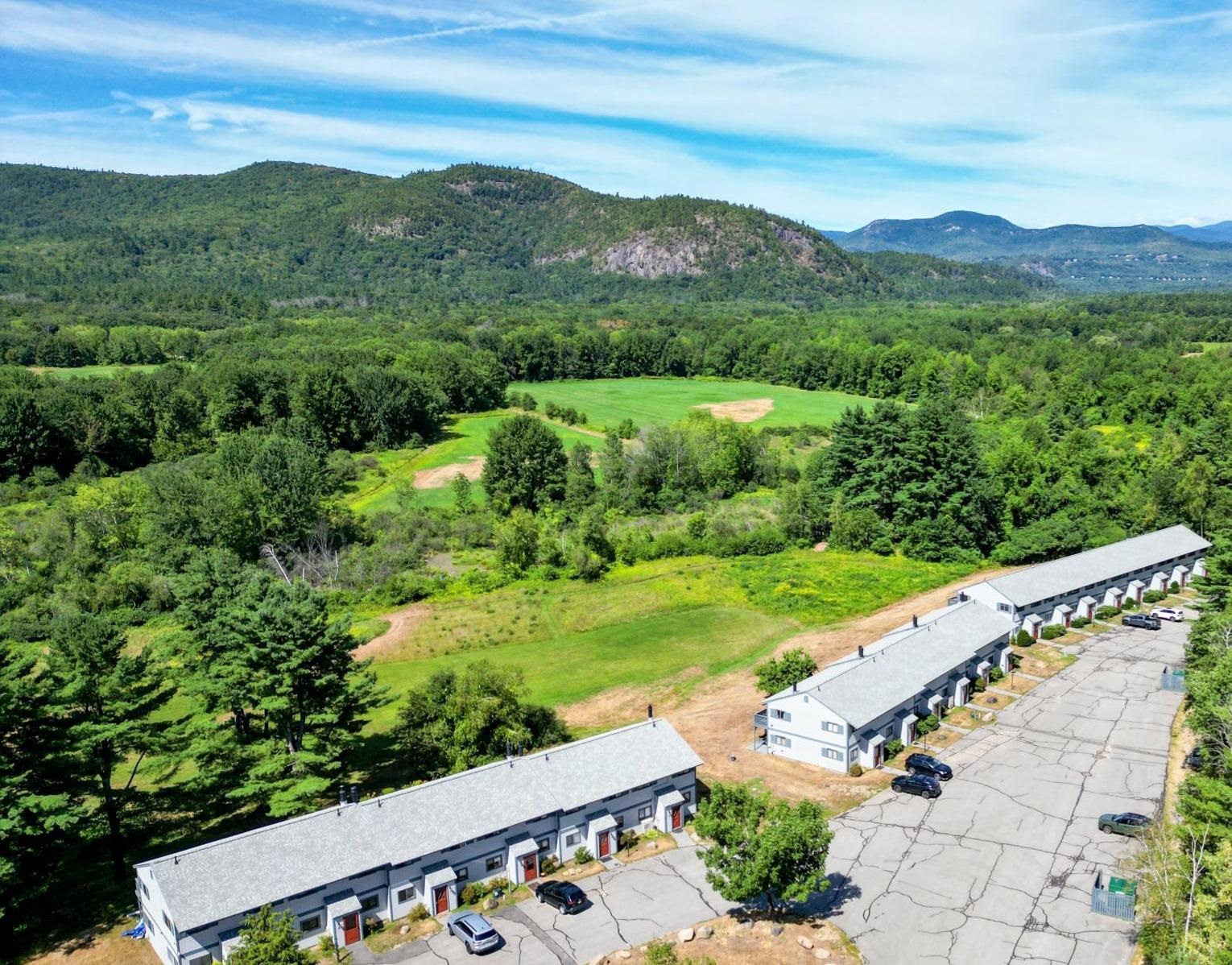 Aerial view of condominium buildings beside a green field, with forested mountains in the distance.