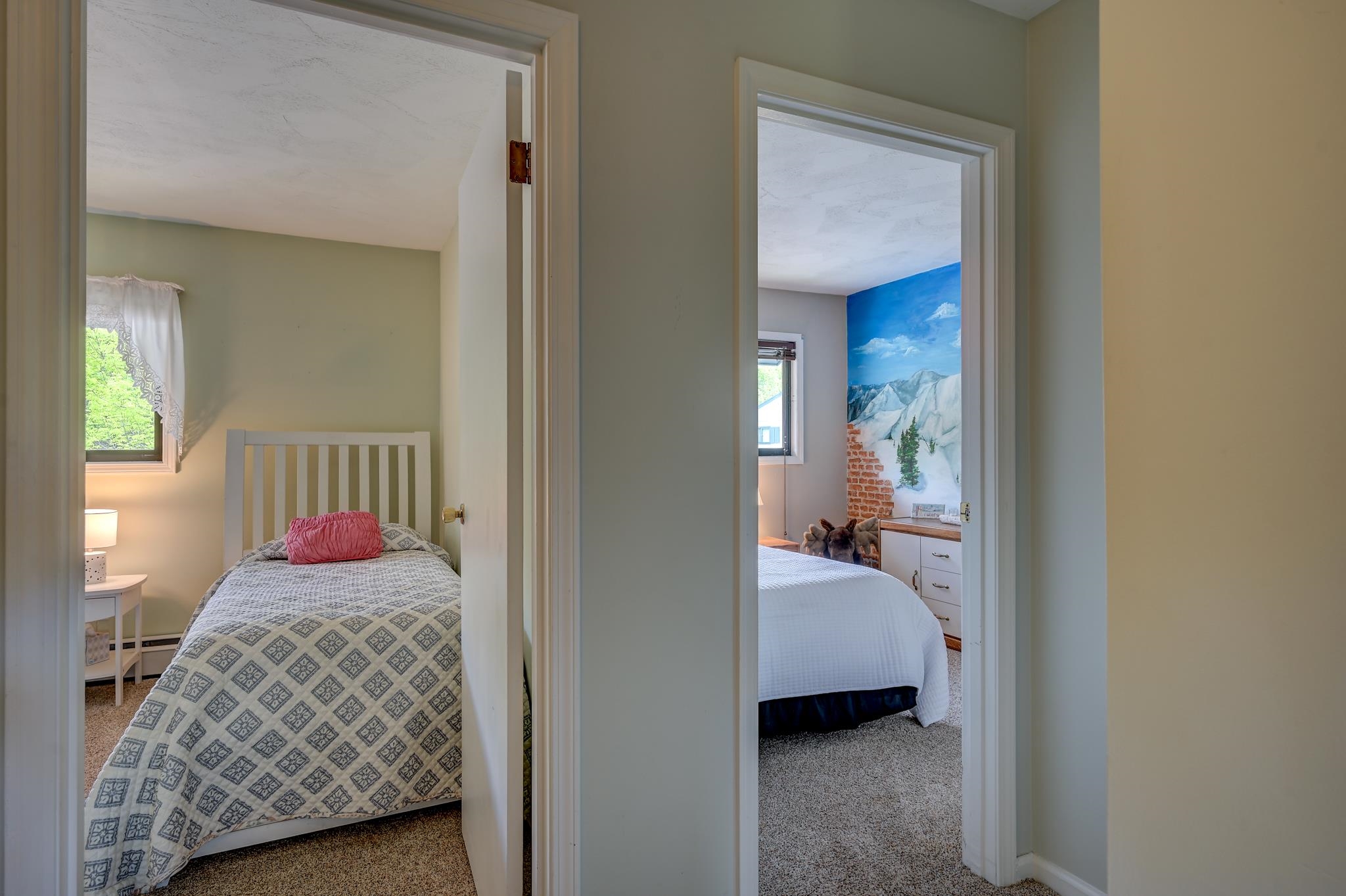 Two furnished bedrooms viewed through open doorways, one with a patterned quilt and the other with a mountain mural.