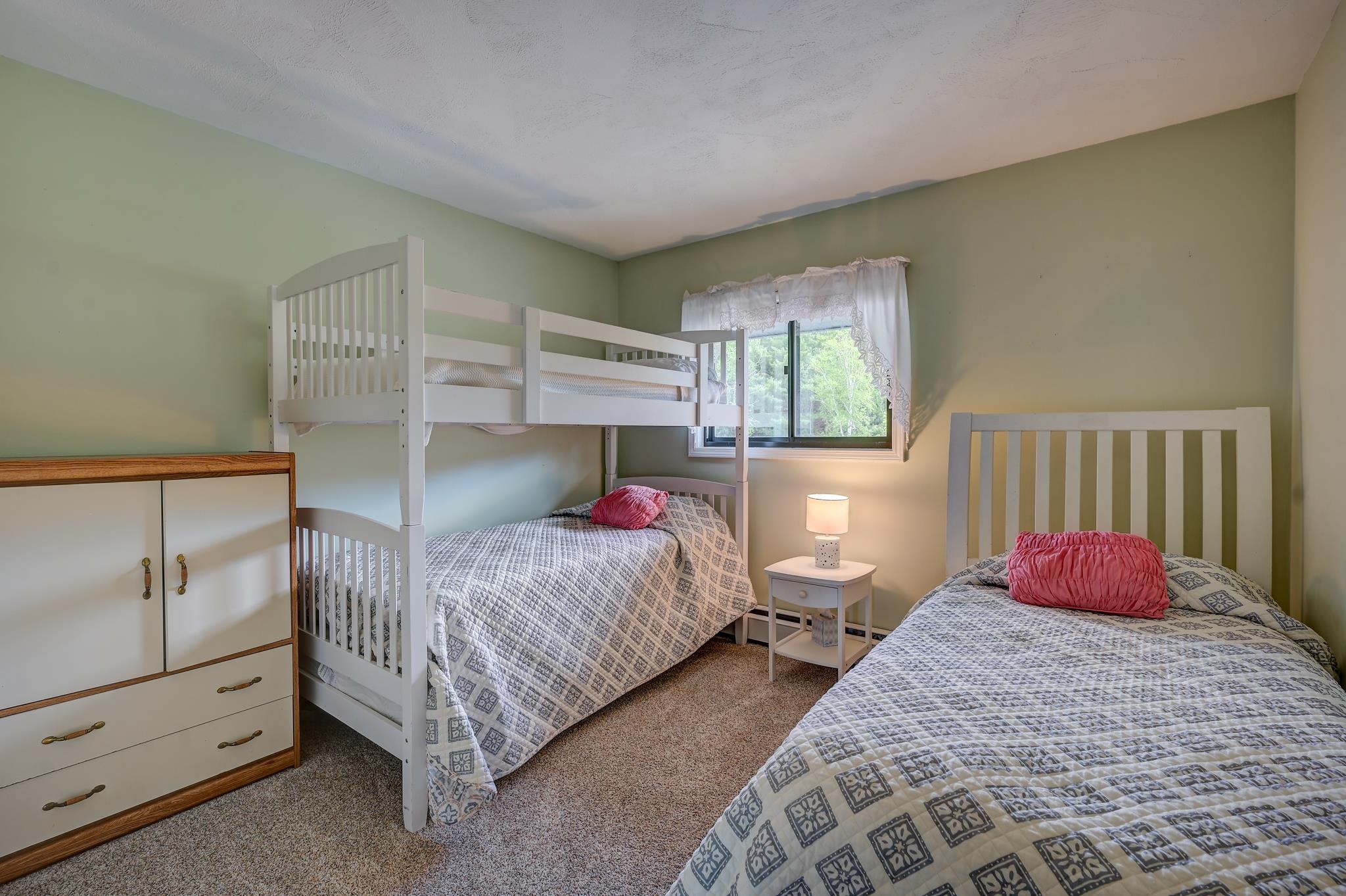 Bedroom with white bunk bed and single bed, patterned quilts, pink pillows, and a window overlooking greenery.