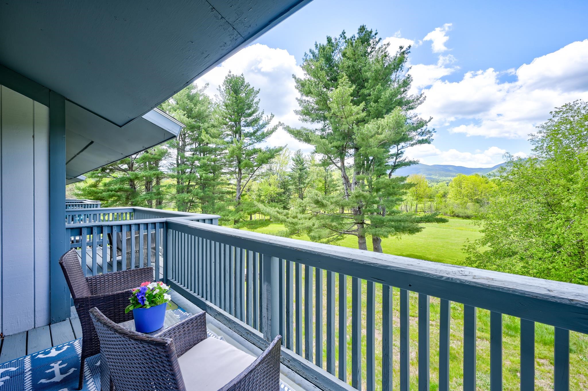Furnished blue-railed balcony overlooking a green lawn, pine trees, and distant mountains.
