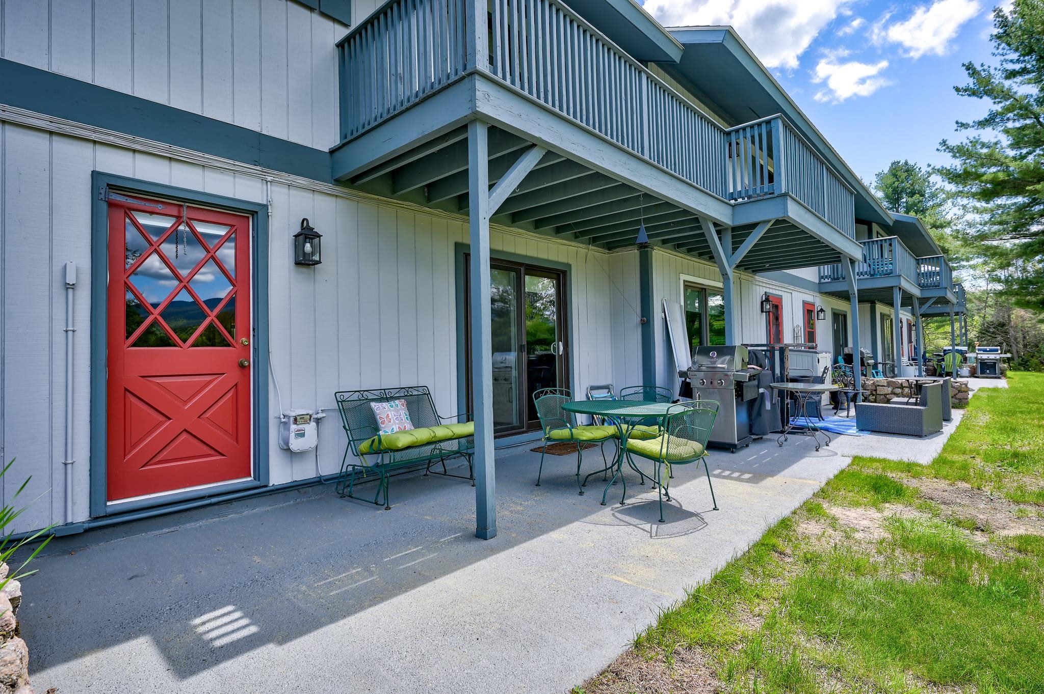 Covered patio with green seating and barbecue beside a white multi-unit building with a red door and balconies.
