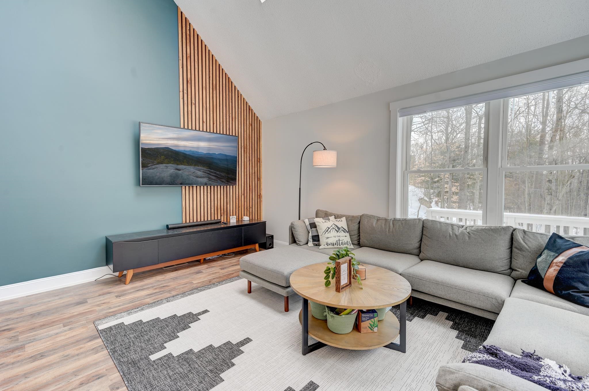 Living room with a gray sectional, round coffee table, blue accent wall, and large windows overlooking snowy trees.