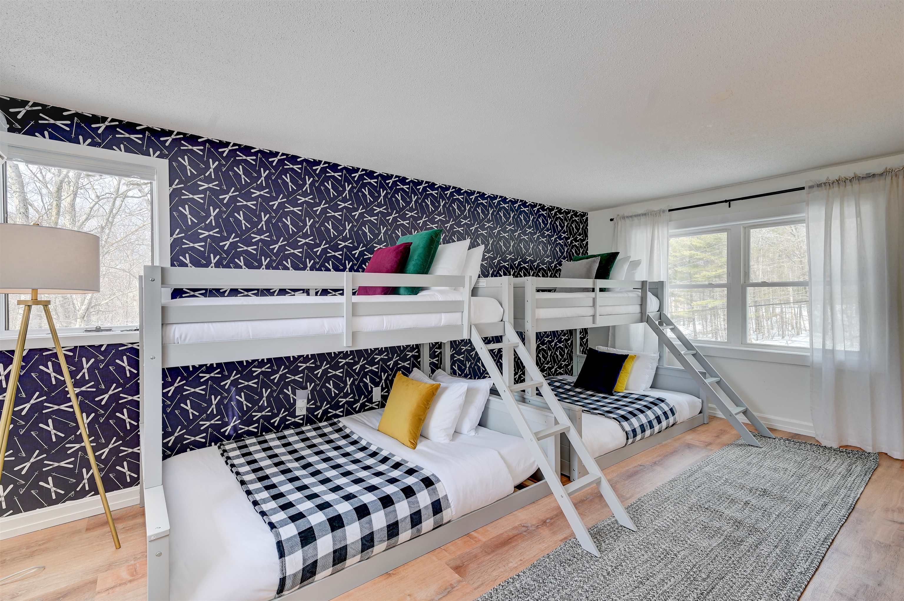 Bunk bedroom with white beds, ski-pattern wallpaper, colorful pillows, and large windows.