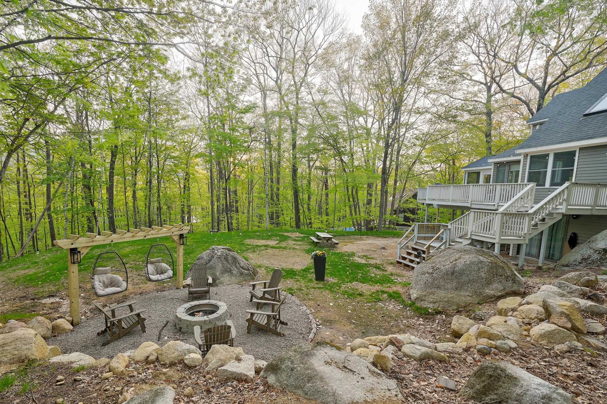 Wooded backyard with multi-level decks, a pergola with swings, and a gravel fire pit seating area.